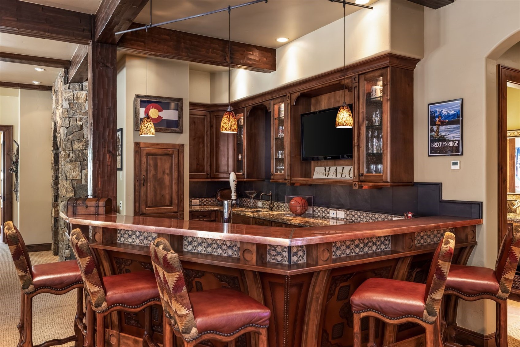 This rustic-style home bar features a prominent copper-topped counter with decorative patterned tile accents and four leather-upholstered bar stools. The space is anchored by rich wood cabinetry, a built-in television, and warm pendant lighting, creating an inviting atmosphere for entertaining. A stone-clad wall adds texture to the room, while framed artwork and a beer tap complete the cozy, lodge-inspired aesthetic.