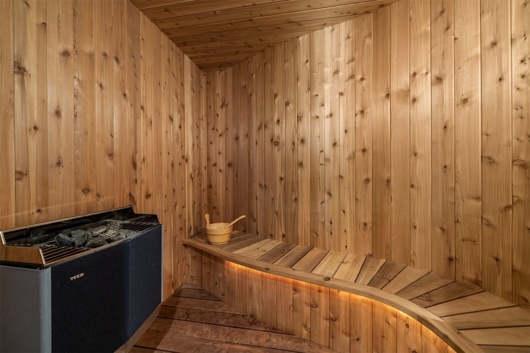 This image showcases a custom-built sauna featuring warm-toned wood paneling throughout the walls, ceiling, and curved bench seating. A modern electric sauna heater with volcanic stones sits in the corner, while a small wooden bucket rests on the bench, which is accented by integrated under-bench lighting. The space offers a serene and high-end wellness experience, characterized by its clean lines and natural material palette.