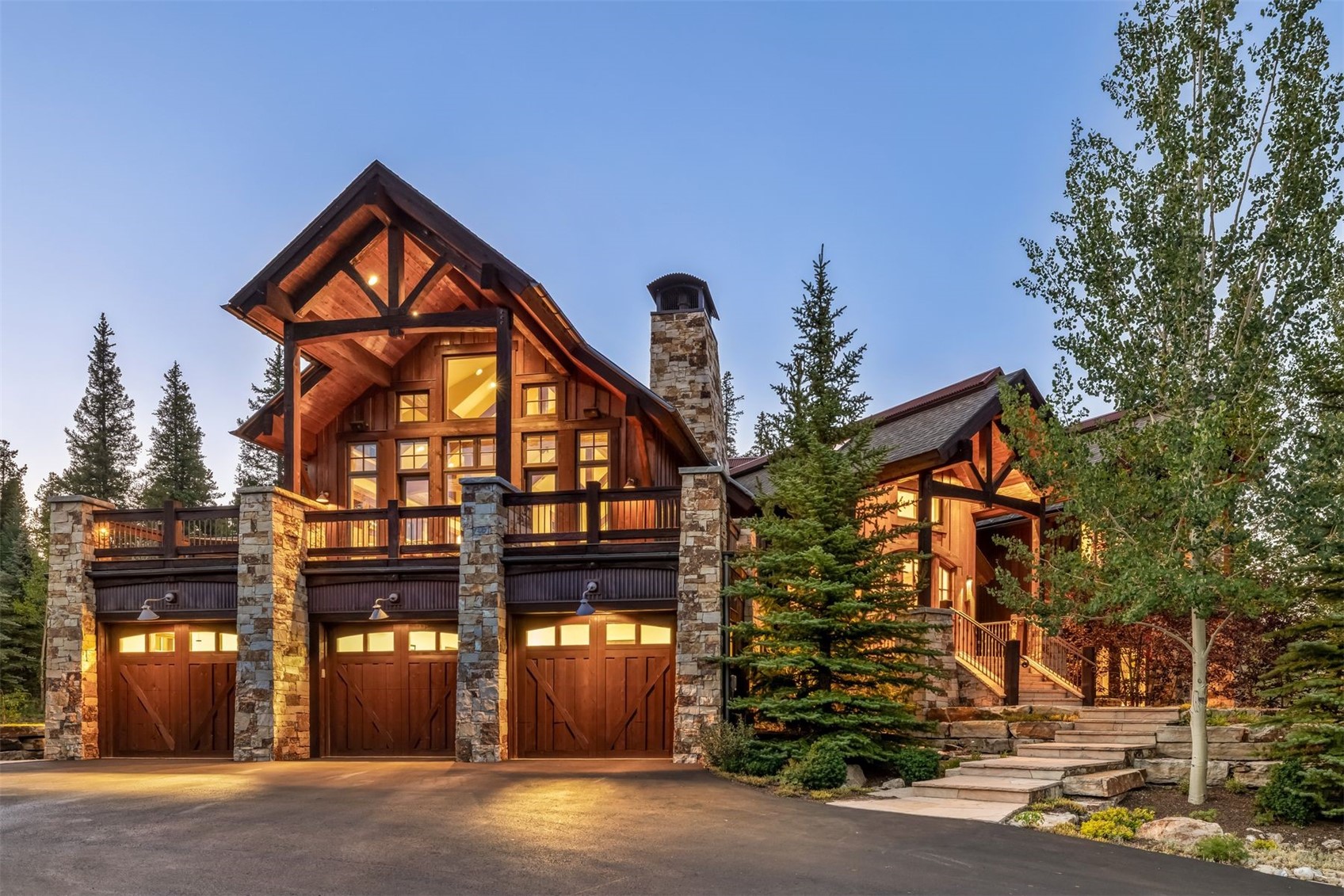 This impressive mountain-style residence showcases a rustic aesthetic with a combination of timber framing, dark wood siding, and extensive stone masonry. The front facade features a prominent three-car garage on the lower level, topped by a spacious balcony and a soaring gabled roofline. A stone-clad chimney breast rises prominently against the structure, while a stone staircase leads to the main entrance, all set against a backdrop of mature pine trees at dusk.