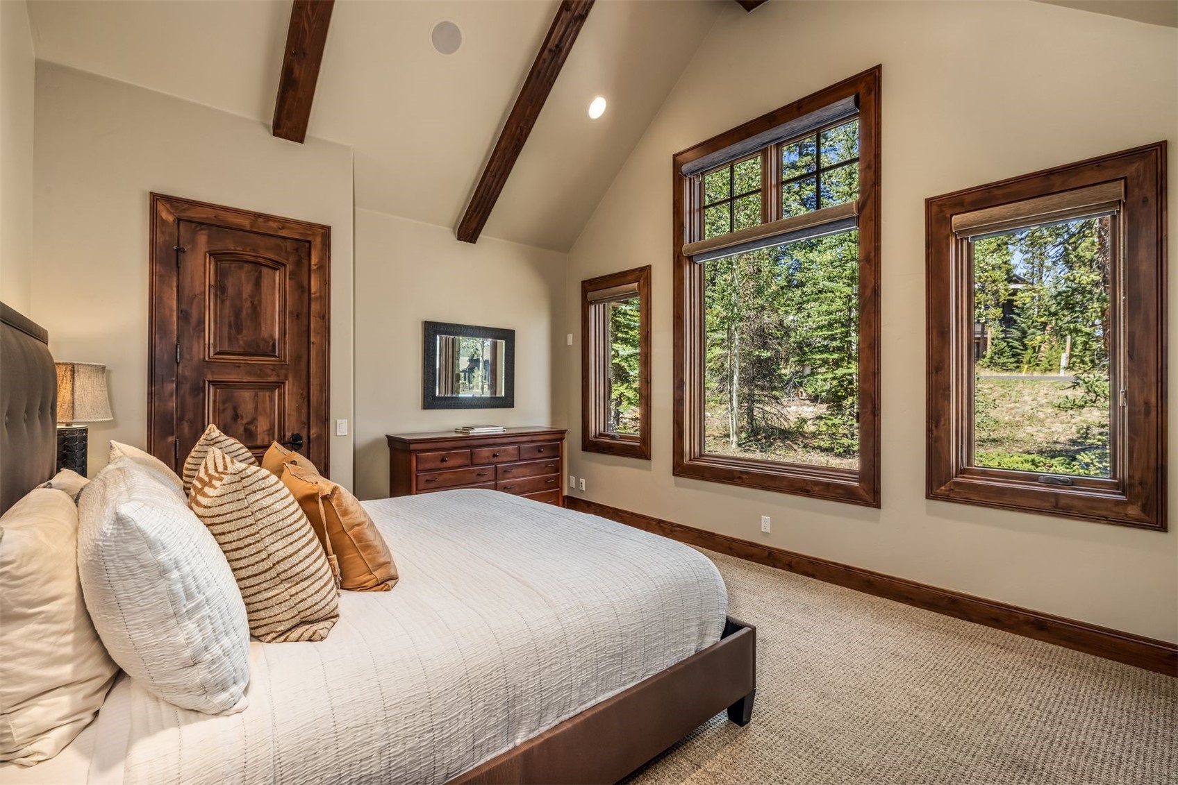 This primary bedroom features a vaulted ceiling with exposed wooden beams and neutral-toned walls, creating a warm and inviting atmosphere. A large bed with textured pillows sits adjacent to a wooden dresser and a mirror, while multiple windows provide ample natural light and views of the surrounding forest. The room is finished with neutral carpeting and rich wood trim around the doors and windows, emphasizing a rustic yet refined aesthetic.