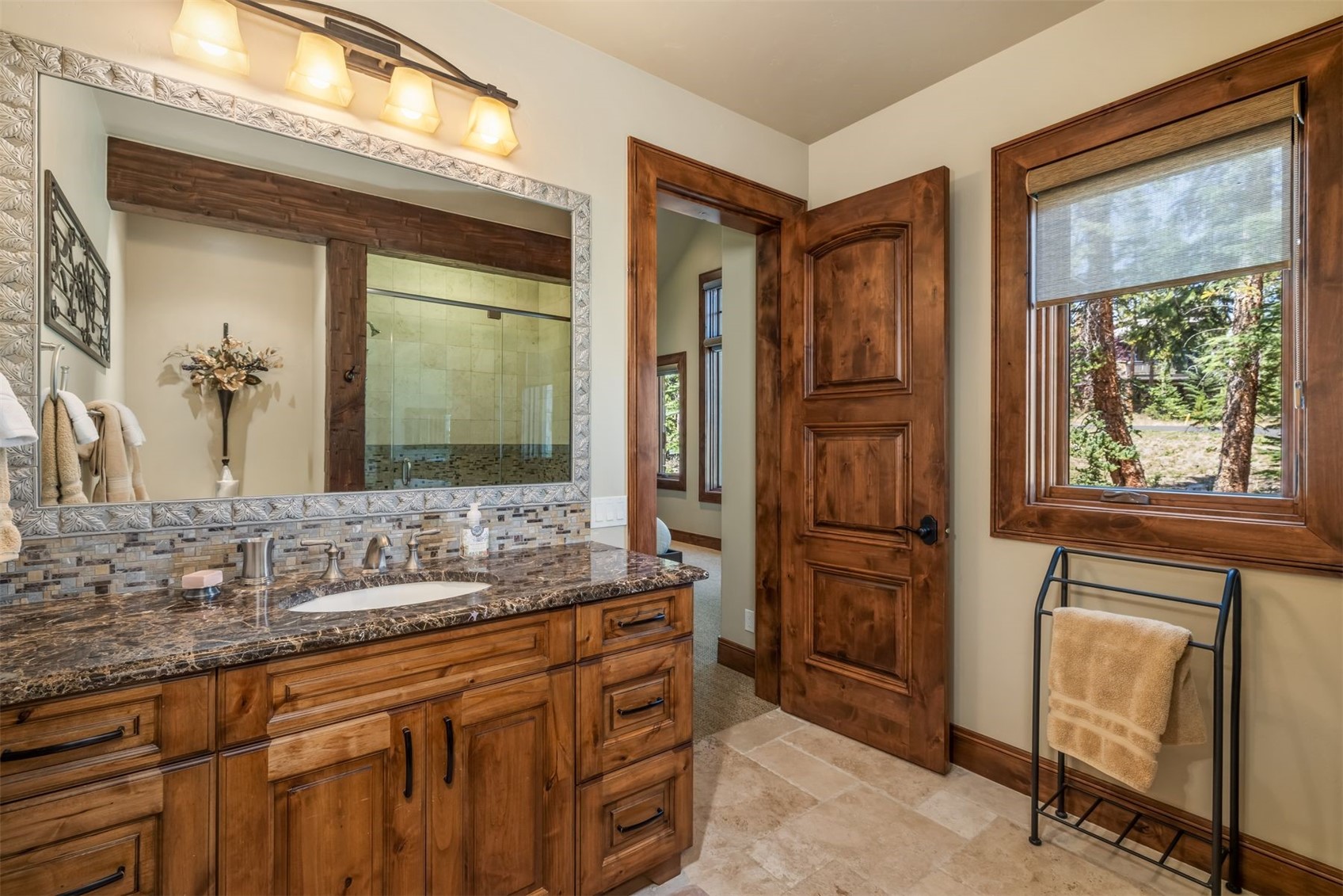 This rustic-style bathroom features a dark stone-topped vanity with warm-toned wood cabinetry and a large, decorative framed mirror. The space includes a tiled backsplash, a wooden door leading to an adjacent room, and a window with a view of the outdoors. A metal towel rack stands near the window, and the room is finished with neutral-toned floor tiles.