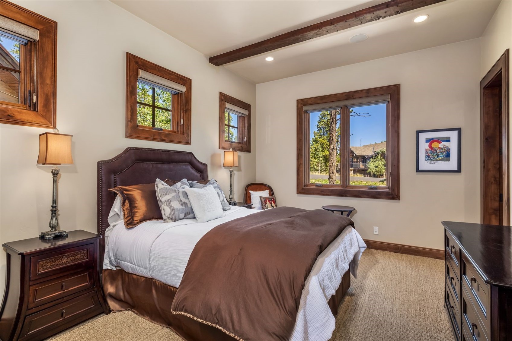This cozy bedroom features a dark leather-upholstered headboard, a plush bed with brown and white bedding, and matching dark wood nightstands with traditional lamps. The room is accented by warm-toned wood window frames, exposed ceiling beams, and a framed piece of art on the wall. The perspective is a straight-on shot from the foot of the bed, highlighting the comfortable and rustic atmosphere of the space.