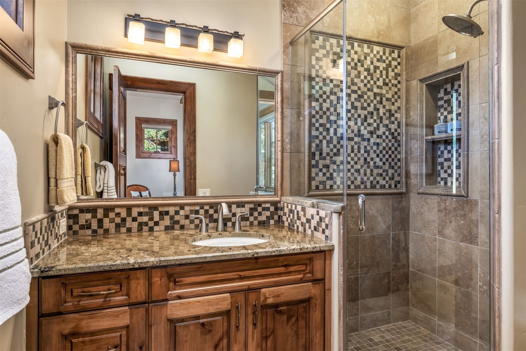 This bathroom features a warm, rustic aesthetic with wood cabinetry, a granite countertop, and a decorative mosaic tile backsplash. A glass-enclosed shower stall sits adjacent to the vanity, showcasing matching stone wall tiles and a built-in recessed niche. The perspective captures the vanity and shower area, highlighting the cohesive design and quality finishes.