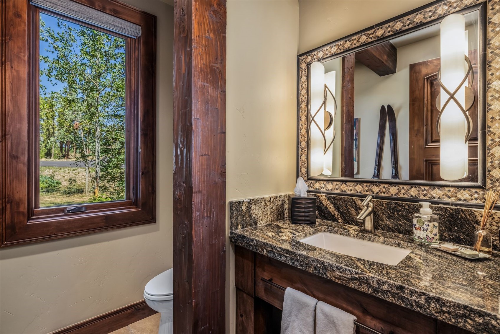 This rustic-style bathroom features a dark wood vanity topped with a patterned stone countertop and an integrated sink. A large mirror with a decorative mosaic tile frame is flanked by two tall, cylindrical wall sconces, while a large window to the left provides natural light and a view of the surrounding trees. A heavy, dark wood structural post separates the vanity area from the toilet, enhancing the cabin-inspired aesthetic.