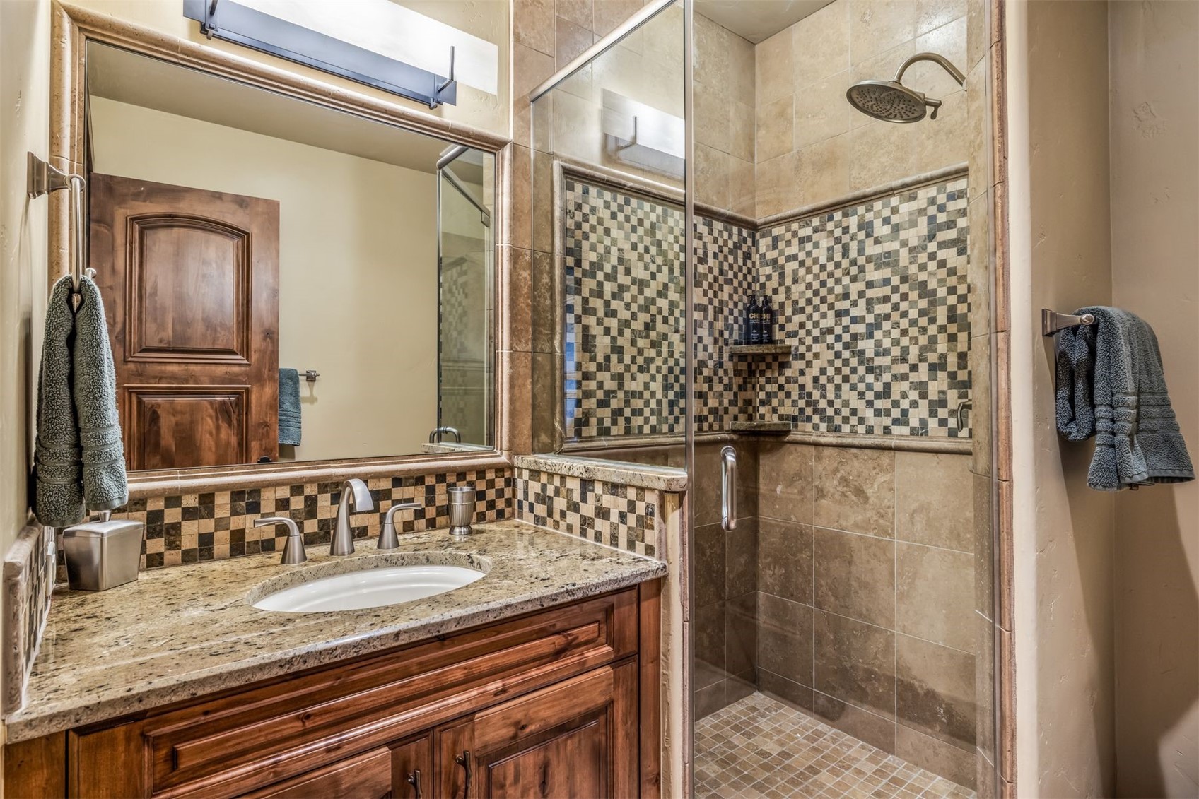 This bathroom features a rustic aesthetic with a wood vanity topped by a stone countertop and an oval undermount sink. A glass-enclosed shower stall is integrated into the space, showcasing a decorative checkered tile accent wall and a rainfall showerhead. The warm, earthy tones of the tile work and the wood cabinetry create a cohesive and inviting atmosphere.