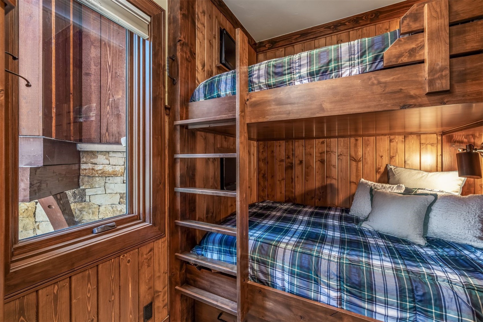This cozy, rustic-style bunk room features custom-built wooden bunk beds with a classic plaid bedding set. The walls are finished with warm-toned wood paneling, and a large window to the left provides a view of an exterior stone wall and wooden structure. The space is intimate and well-lit, creating a comfortable and inviting atmosphere typical of a mountain cabin or lodge.