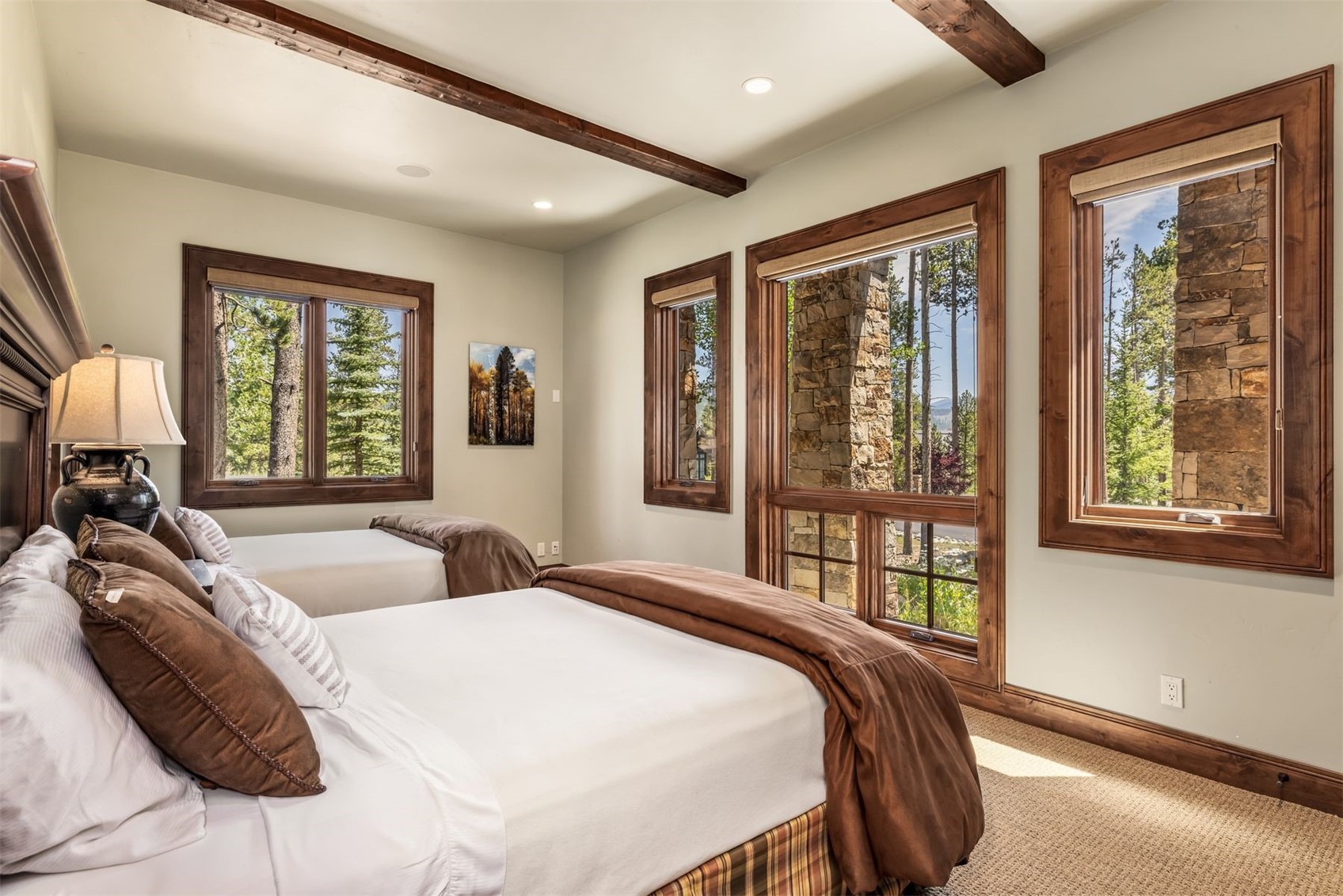 This guest bedroom features two beds with brown bedding and a neutral color palette, accented by dark wood trim around the windows and ceiling beams. Large windows provide ample natural light and views of the surrounding forest and stone exterior elements. The room has a cozy, rustic atmosphere with carpeted flooring and a bedside lamp providing warm lighting.