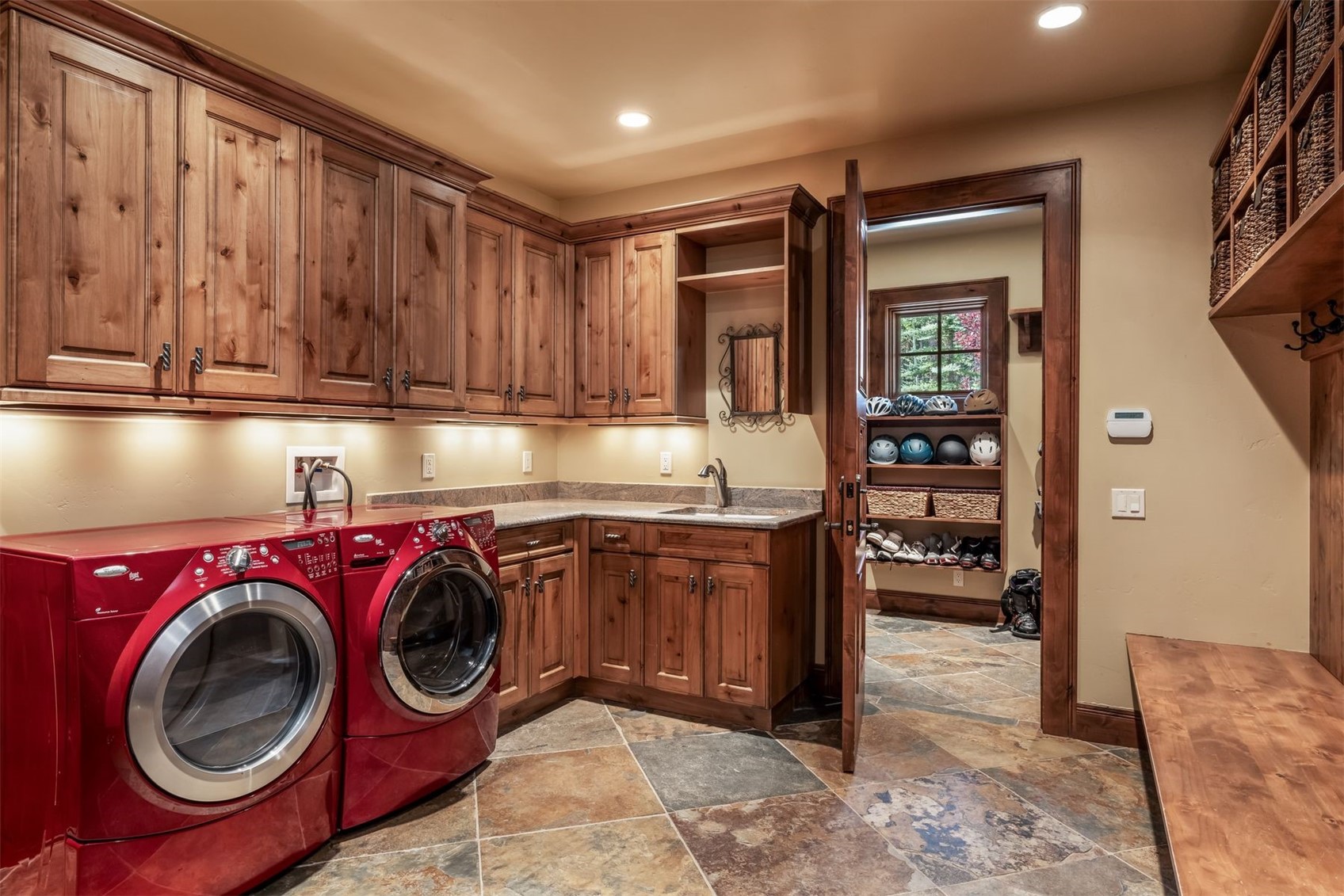 This functional laundry room features vibrant red front-loading appliances set against warm-toned wood cabinetry and stone-look tile flooring. The space includes a built-in sink, under-cabinet lighting, and an open doorway leading to a mudroom area equipped with shelving for helmets and footwear. The overall aesthetic is rustic and organized, providing a practical utility space with ample storage.