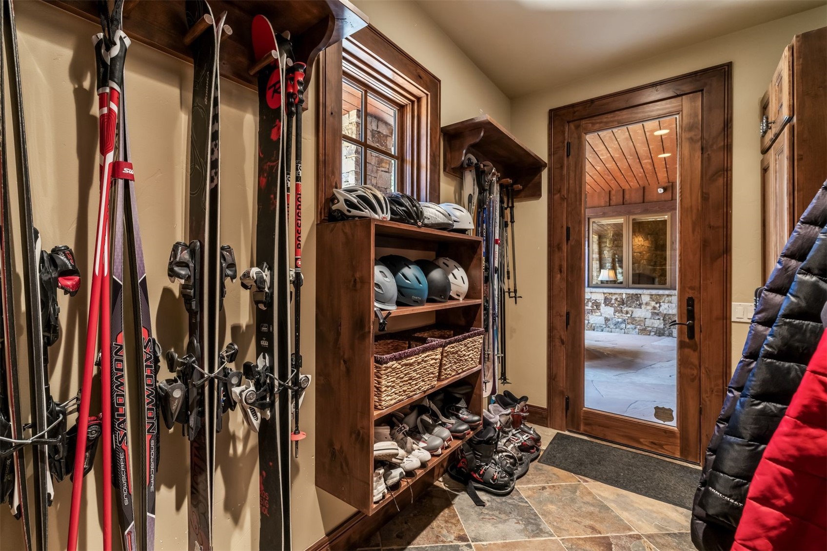 This mudroom or ski storage area features a rustic aesthetic with warm wood trim and stone-look tile flooring. The space is efficiently organized with wall-mounted ski racks, a built-in shelving unit for helmets and boots, and a glass-paneled door that provides a view into an adjacent stone-walled room. The warm lighting and functional storage layout create a welcoming and practical transition space for outdoor enthusiasts.