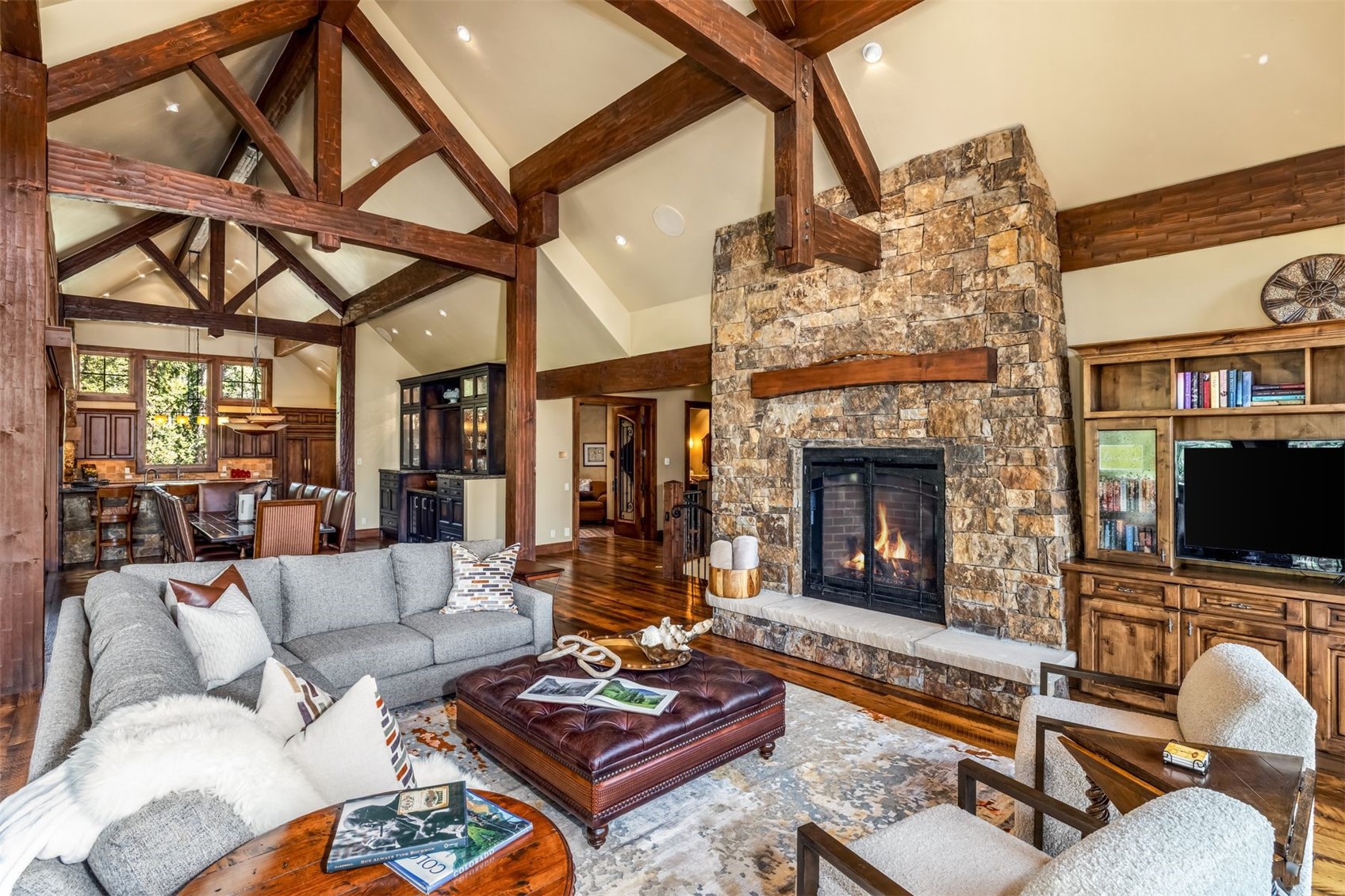 This spacious living room features a vaulted ceiling with exposed timber trusses and a prominent stone-clad chimney breast housing a fireplace. The open-concept layout flows into a dining area and kitchen, showcasing warm wood tones and a rustic, mountain-inspired aesthetic. A large gray sectional sofa and a leather ottoman provide comfortable seating, while the room is anchored by a patterned area rug and built-in cabinetry housing a television.