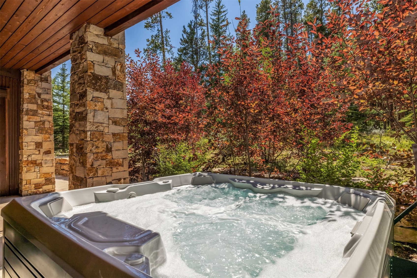 This image captures a relaxing outdoor spa experience set on a covered patio. A bubbling hot tub with molded seating is the focal point, positioned next to a sturdy stone pillar that supports the wooden ceiling structure. Beyond the patio, vibrant red-leafed trees and evergreen pines create a natural, secluded backdrop, offering a serene and private atmosphere.