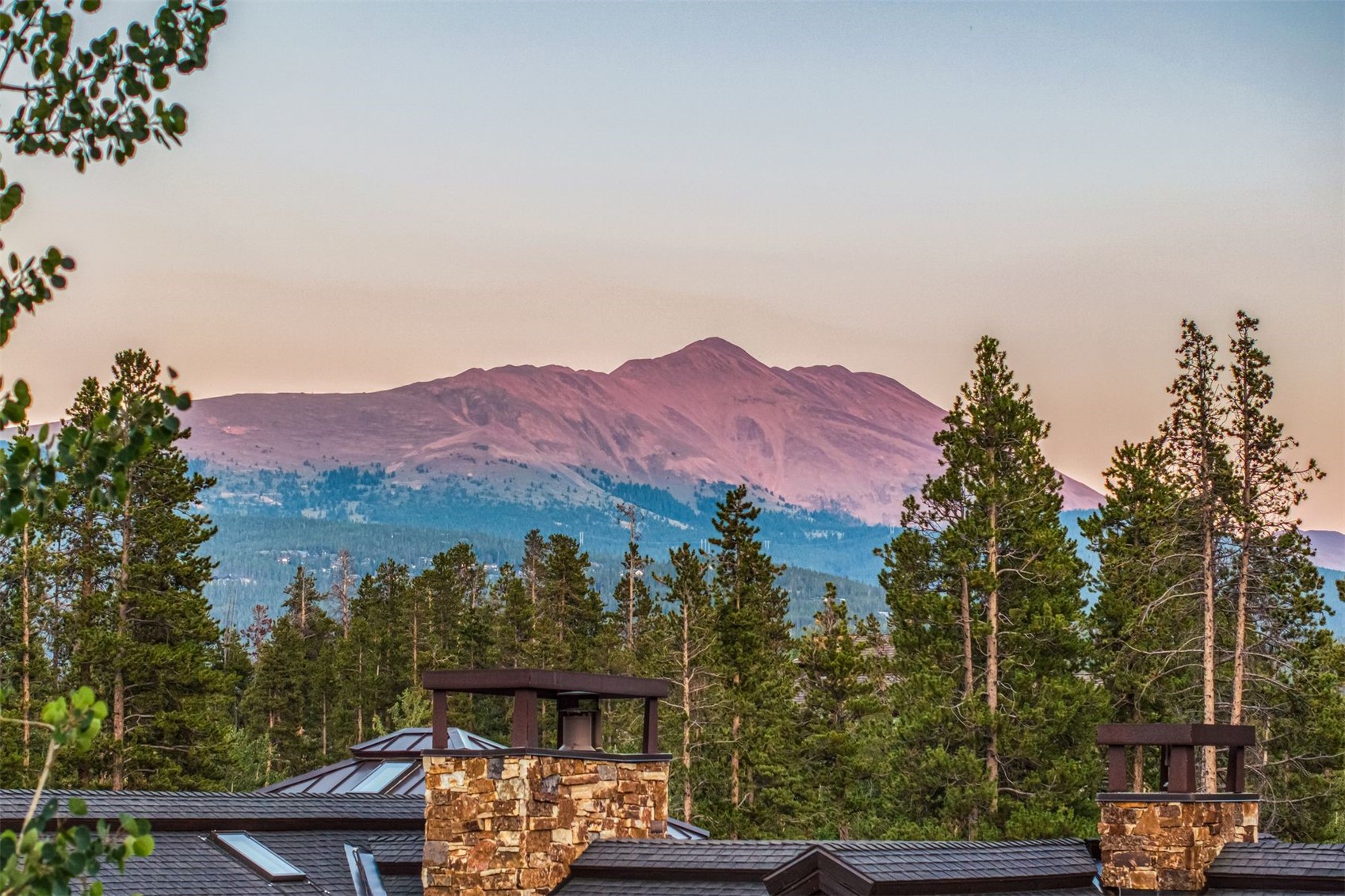 This elevated perspective captures the rear roofline of a mountain residence, showcasing dark shingled roofing and prominent stone-clad chimneys. The foreground features a dense canopy of evergreen trees, while the background is dominated by a majestic, sun-drenched mountain peak bathed in the soft, pink hues of sunset. The composition emphasizes the property's integration into a serene, high-altitude natural landscape.