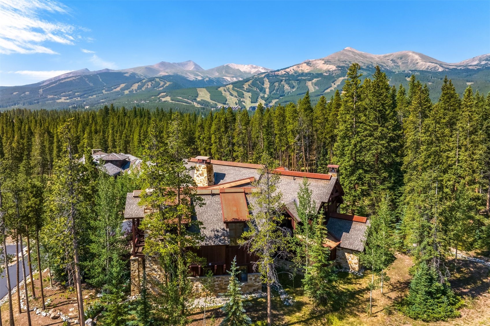 This high-angle aerial shot captures a sprawling mountain lodge nestled within a dense forest of evergreen trees. The home features a complex, multi-gabled roofline with metal accents and stone exterior walls, perfectly blending into the rugged, high-altitude landscape. In the background, a majestic mountain range with visible ski runs provides a dramatic and scenic backdrop for this secluded retreat.