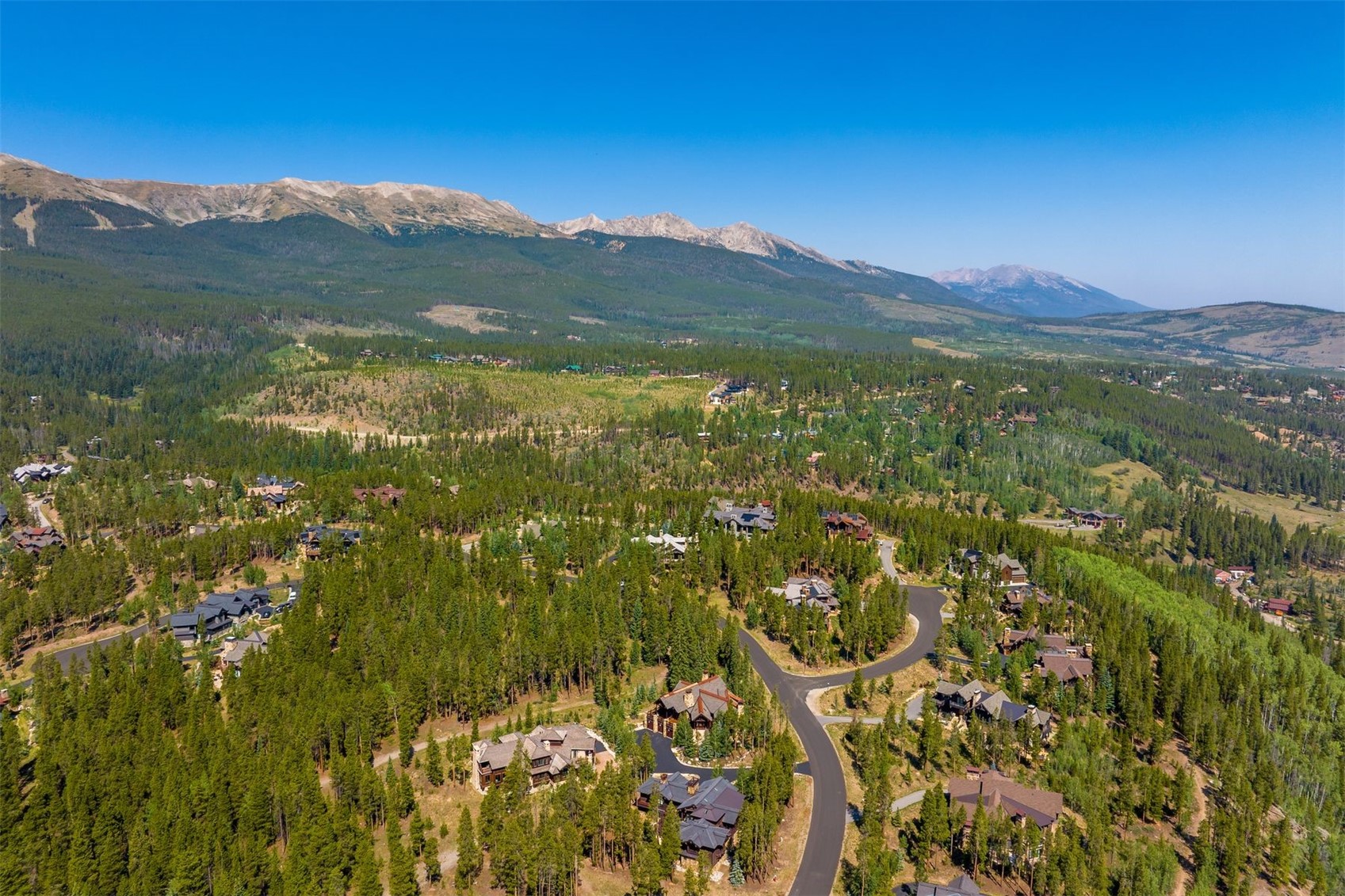 This high-angle aerial perspective captures a sprawling mountain community nestled within a dense evergreen forest. The scene features several large, multi-story homes with dark-toned roofs and wood-accented exteriors, connected by winding paved roads. In the background, majestic, rugged mountain peaks rise against a clear, expansive blue sky, emphasizing the serene and secluded nature of the location.