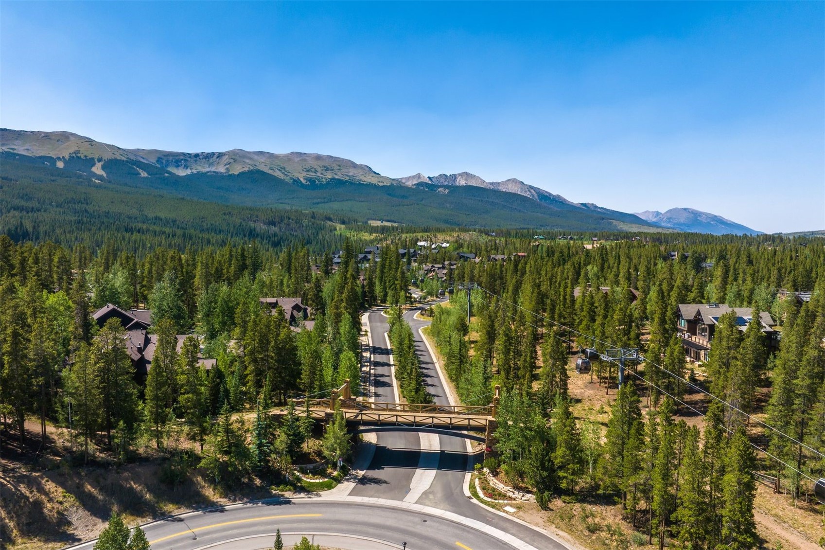 This high-angle aerial shot captures a scenic mountain community featuring a winding road that passes beneath a rustic wooden pedestrian bridge. The landscape is densely forested with evergreen trees, and a gondola lift system traverses the air above the road, highlighting the area's recreational appeal. In the background, majestic mountain peaks rise against a clear blue sky, creating a serene and picturesque setting.