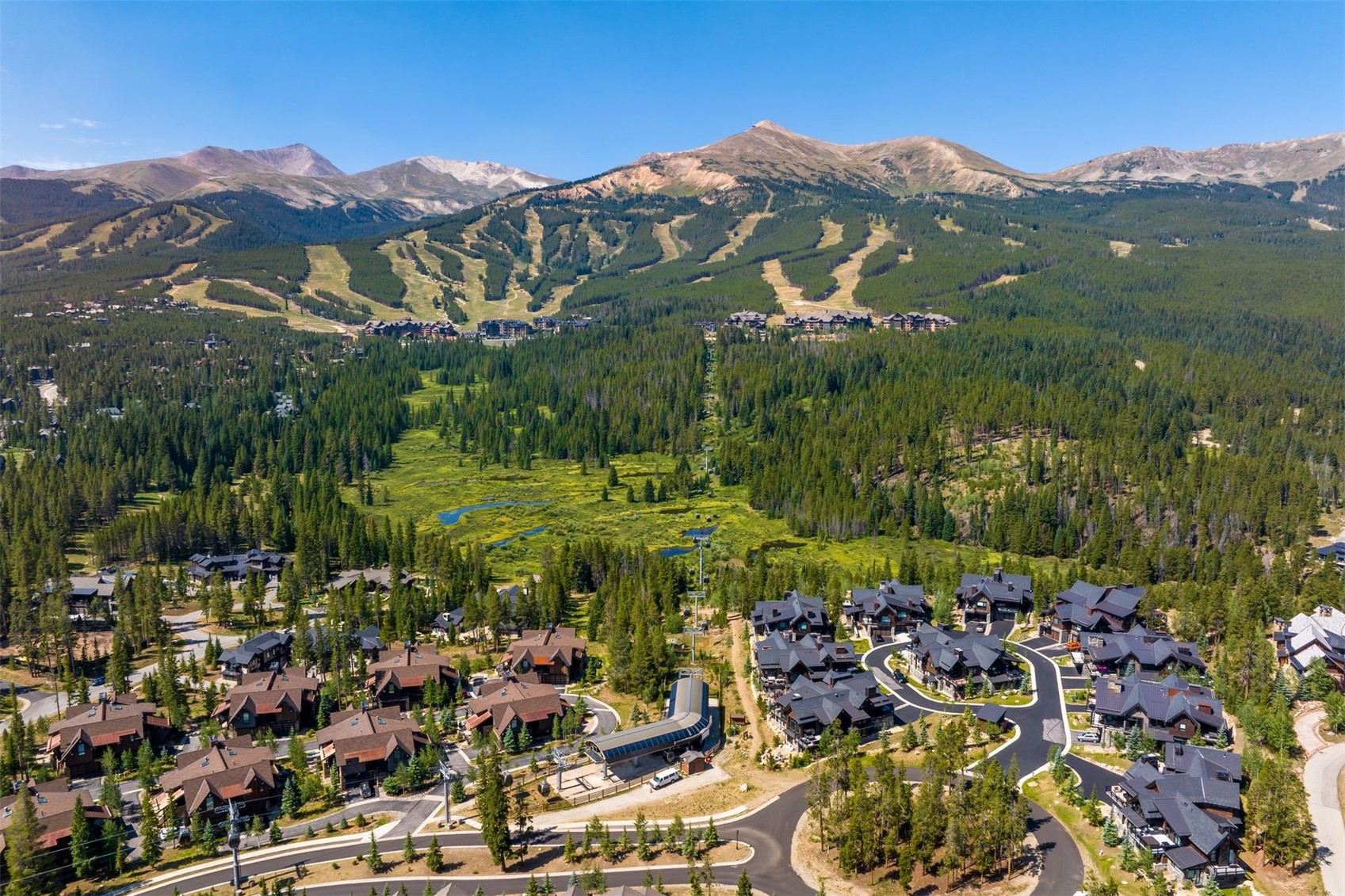 This high-angle aerial view captures a luxury mountain community nestled at the base of a sprawling ski resort. The scene features clusters of multi-story mountain-style homes with dark roofs and wood siding, integrated into a dense evergreen forest. A ski lift station is visible in the foreground, with groomed ski runs cutting through the mountain slopes in the background under a clear blue sky.