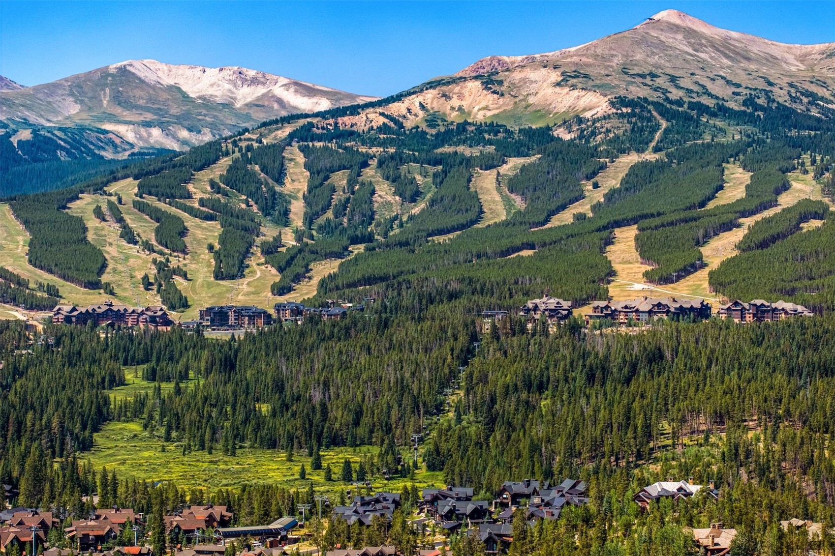 This expansive aerial view captures a mountain resort community nestled at the base of a vast, forested ski slope. The landscape features a mix of dense evergreen trees, winding ski runs, and clusters of multi-story residential buildings with dark-toned roofs. The scene is set against a backdrop of towering, rugged mountain peaks under a clear blue sky, emphasizing the property's prime location for outdoor recreation.