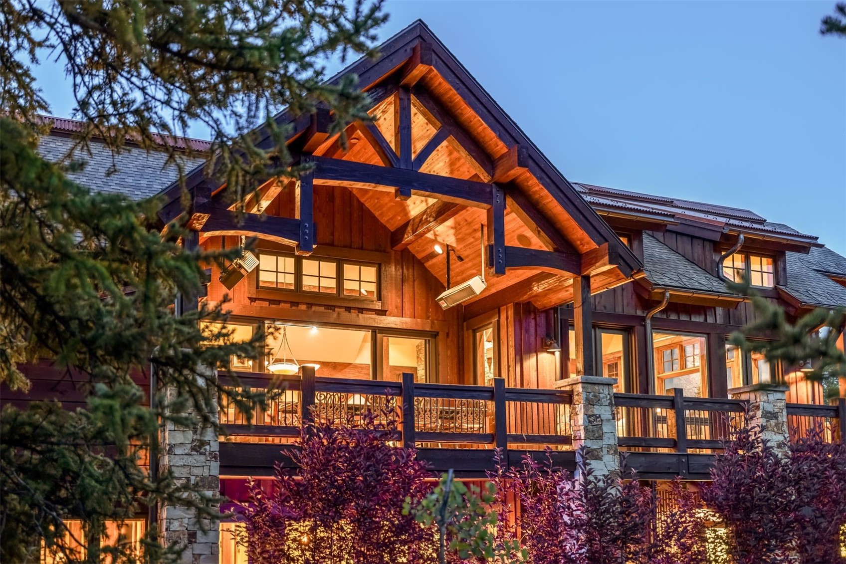 This stunning mountain-style home features a prominent timber-framed gable roof over a spacious, elevated deck. The exterior is clad in rich, dark wood siding accented by natural stone pillars, creating a warm and inviting atmosphere at dusk. Large windows and glass doors provide a seamless transition between the interior living space and the outdoor balcony, which is equipped with a mounted heater for year-round enjoyment.