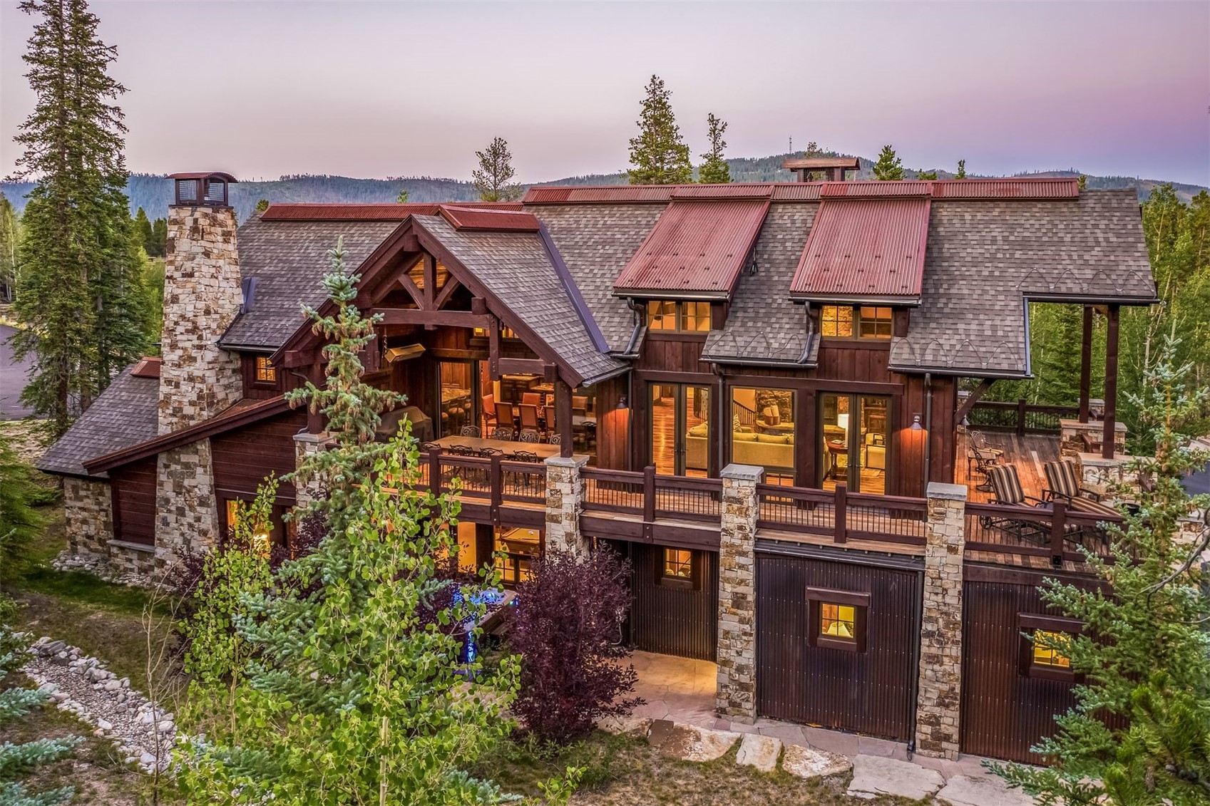 This impressive mountain-style residence features a multi-level design with a blend of dark wood siding, stone accents, and corrugated metal roofing. The rear elevation showcases a spacious elevated deck with outdoor seating, large windows that reveal warm interior lighting, and a prominent stone-clad chimney breast. Set against a backdrop of evergreen trees and a soft twilight sky, the home exudes a luxurious yet rustic aesthetic.