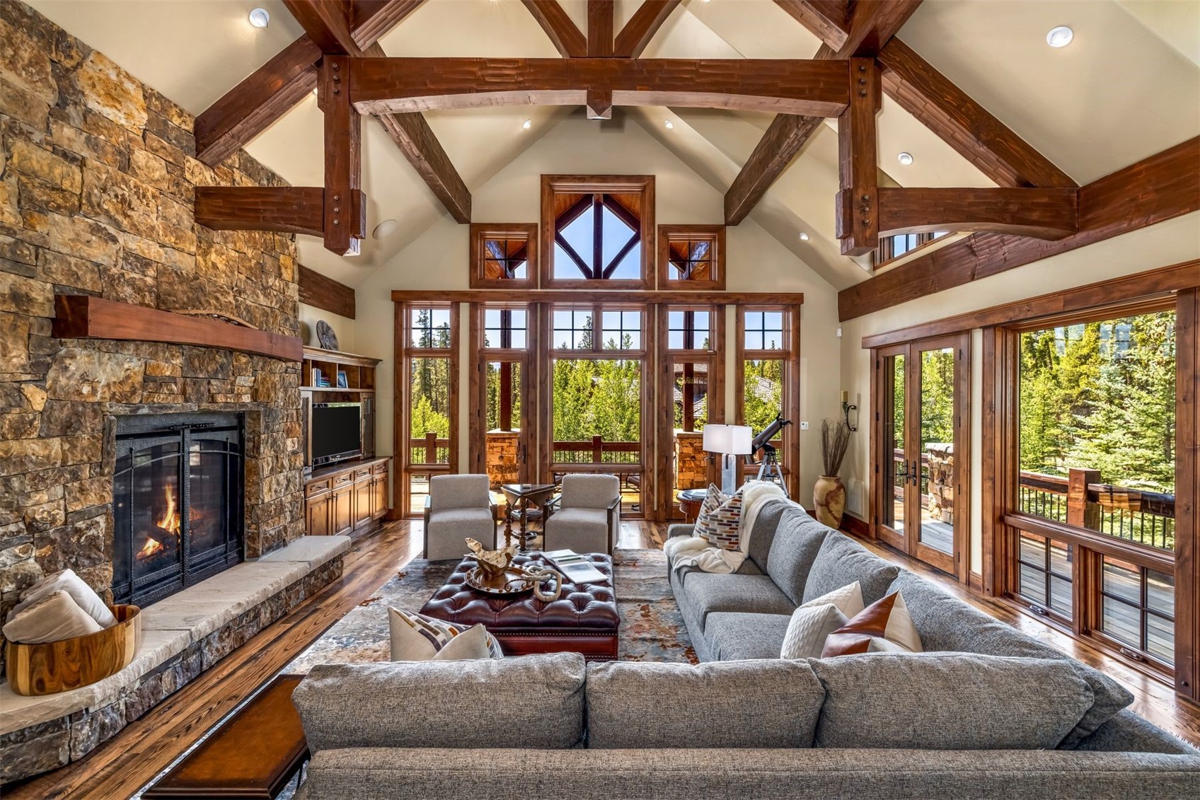 This expansive living room features a vaulted ceiling with exposed timber beams and a prominent stone-clad chimney breast housing a fireplace. A large gray sectional sofa and a leather ottoman are arranged on a patterned rug, facing a wall of tall windows that provide abundant natural light and views of the surrounding trees. The space is defined by its rustic, lodge-inspired aesthetic, characterized by warm wood trim and a cozy, inviting atmosphere.