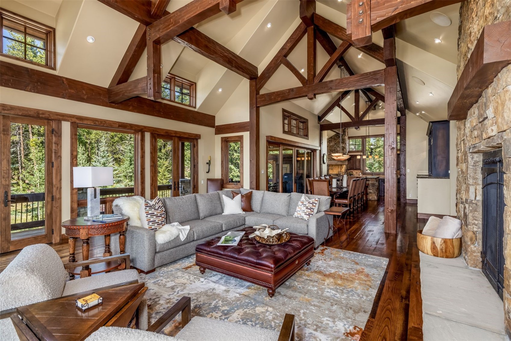This spacious living room features a vaulted ceiling with exposed timber beams and a prominent stone-clad chimney breast. A large gray sectional sofa and a tufted leather ottoman are arranged on a patterned area rug, creating a cozy seating area. The room is bathed in natural light from large windows and glass doors that offer views of the surrounding forest, while a dining area is visible in the background.