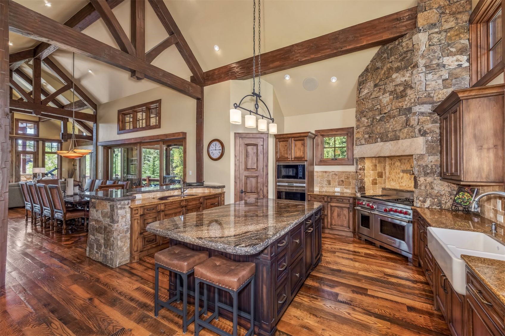 This rustic-style kitchen features a large central island with dark cabinetry and stone countertops, complemented by warm wood flooring and exposed timber beams on the vaulted ceiling. A prominent stone-clad chimney breast houses the range, while a farmhouse-style sink and high-end stainless steel appliances complete the space. The open-concept layout flows seamlessly into an adjacent dining area, creating a spacious and inviting atmosphere.