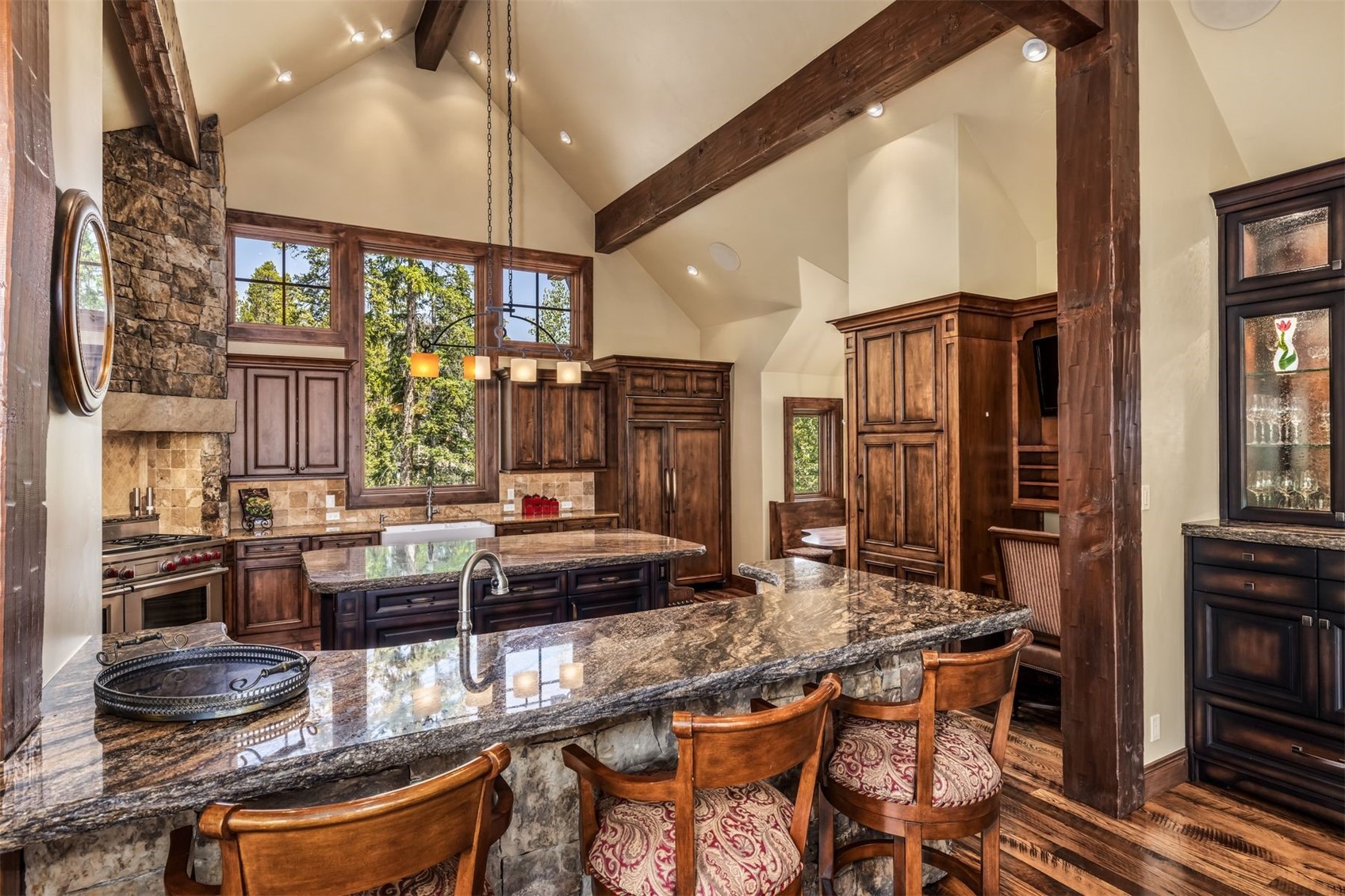 This rustic-style kitchen features dark wood cabinetry, a large island with a stone countertop, and a breakfast bar with three upholstered stools. A stone-clad chimney breast rises above the professional-grade range, while large windows provide ample natural light and views of the surrounding trees. The space is defined by vaulted ceilings with exposed timber beams and a warm, inviting atmosphere.