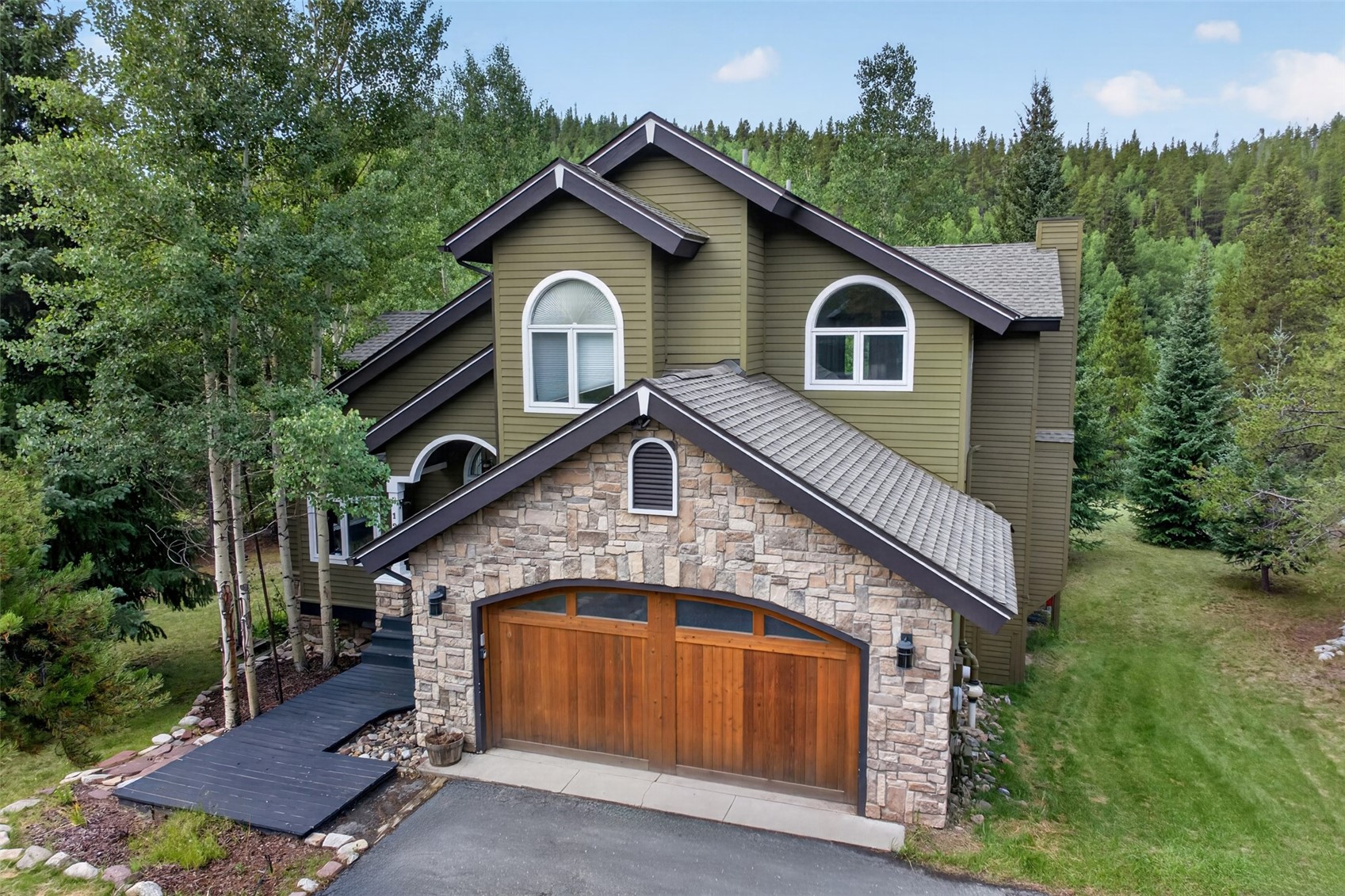 This elevated perspective captures the front facade of a multi-story home nestled in a wooded, mountainous setting. The exterior features a combination of olive-toned horizontal siding and a prominent stone-clad garage section with a large, arched wooden door. The home is framed by tall aspen and pine trees, creating a secluded, rustic atmosphere.