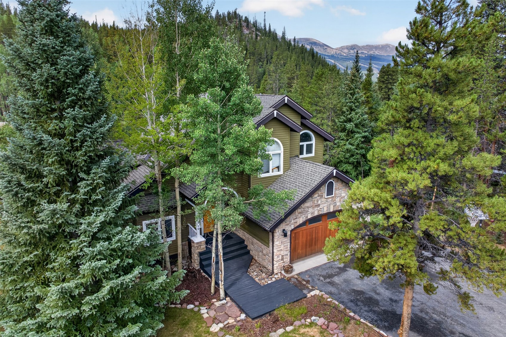 This elevated aerial perspective captures a mountain-style home nestled within a dense forest of evergreen and aspen trees. The house features a multi-gabled roofline, a combination of green siding and stone veneer, and a prominent wooden garage door. A dark-painted wooden walkway leads to the front entrance, creating a striking contrast against the natural landscape and the rugged mountain backdrop.