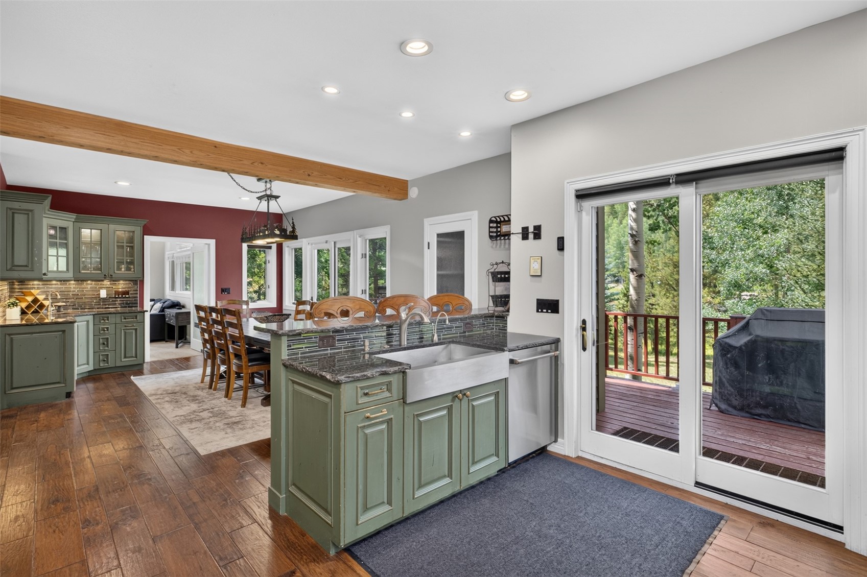 This open-concept kitchen features sage green cabinetry paired with dark stone countertops and a farmhouse-style sink. A wooden ceiling beam adds a rustic touch, while the space flows seamlessly into a dining area and opens to an outdoor deck through sliding glass doors. The warm wood flooring and contrasting red accent wall in the dining area create a cozy, inviting atmosphere.