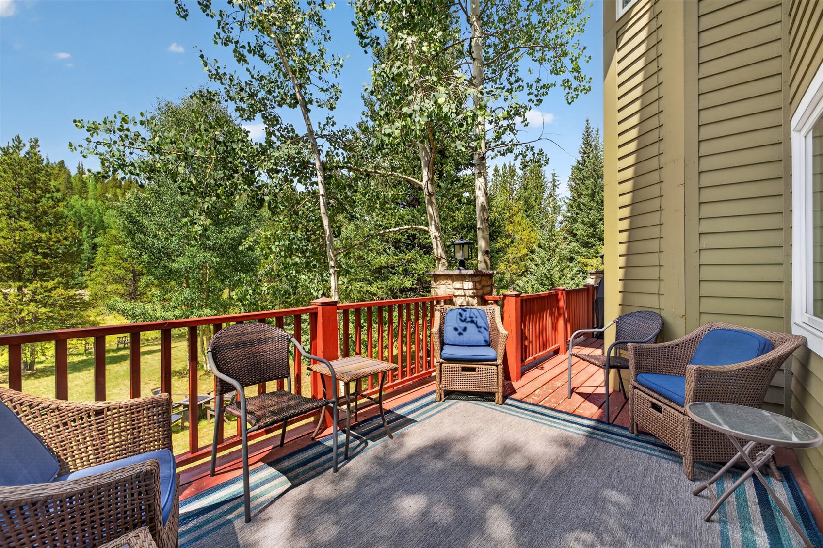 This elevated wooden deck offers a serene outdoor living space furnished with several wicker-style chairs and small side tables arranged on a patterned area rug. The deck is enclosed by a red railing and overlooks a lush, wooded landscape filled with tall aspen and pine trees. The perspective captures a peaceful, sunny afternoon setting, highlighting the home's connection to the surrounding natural environment.