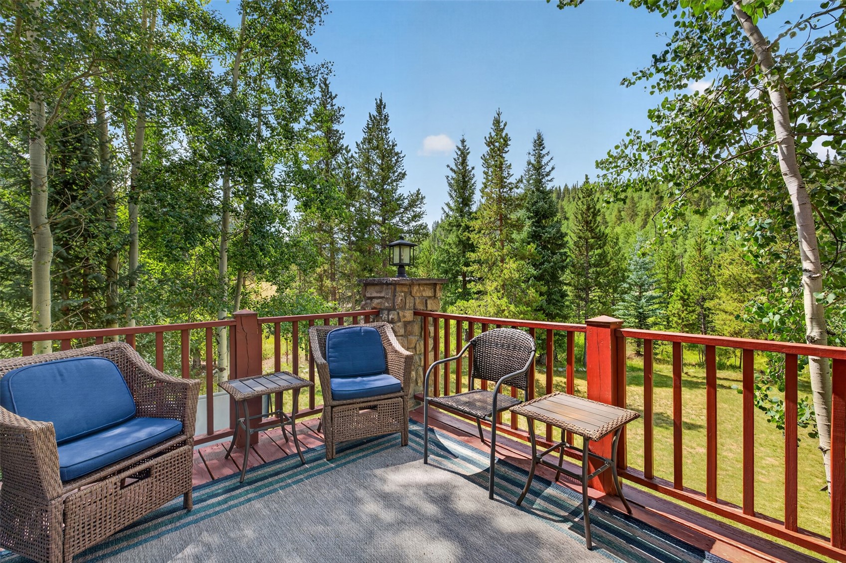 This elevated wooden deck offers a serene outdoor living space furnished with two wicker-style armchairs with blue cushions and two matching side tables. The deck is enclosed by a red-painted railing and features a stone-clad pillar topped with a lantern-style light fixture. Beyond the railing, a lush, tree-filled landscape provides a private and tranquil natural backdrop.