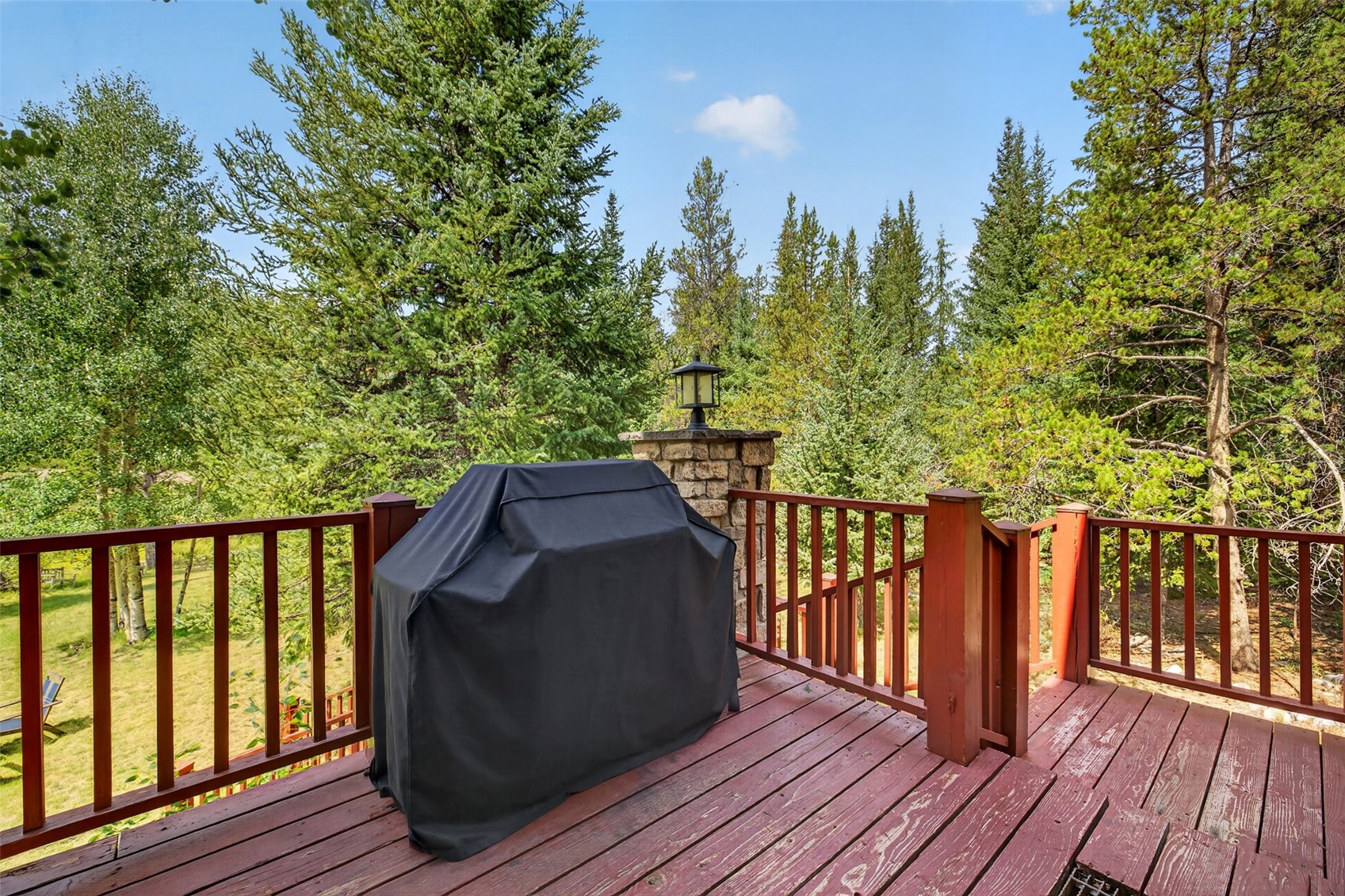This elevated wooden deck offers a serene outdoor space surrounded by a dense forest of pine and aspen trees. A grill covered by a black protective tarp sits prominently on the deck, which is enclosed by a stained wood railing and features a stone-clad pillar topped with an outdoor light fixture. The perspective captures the deck from a slightly elevated angle, emphasizing the peaceful, wooded setting and the potential for outdoor dining or relaxation.