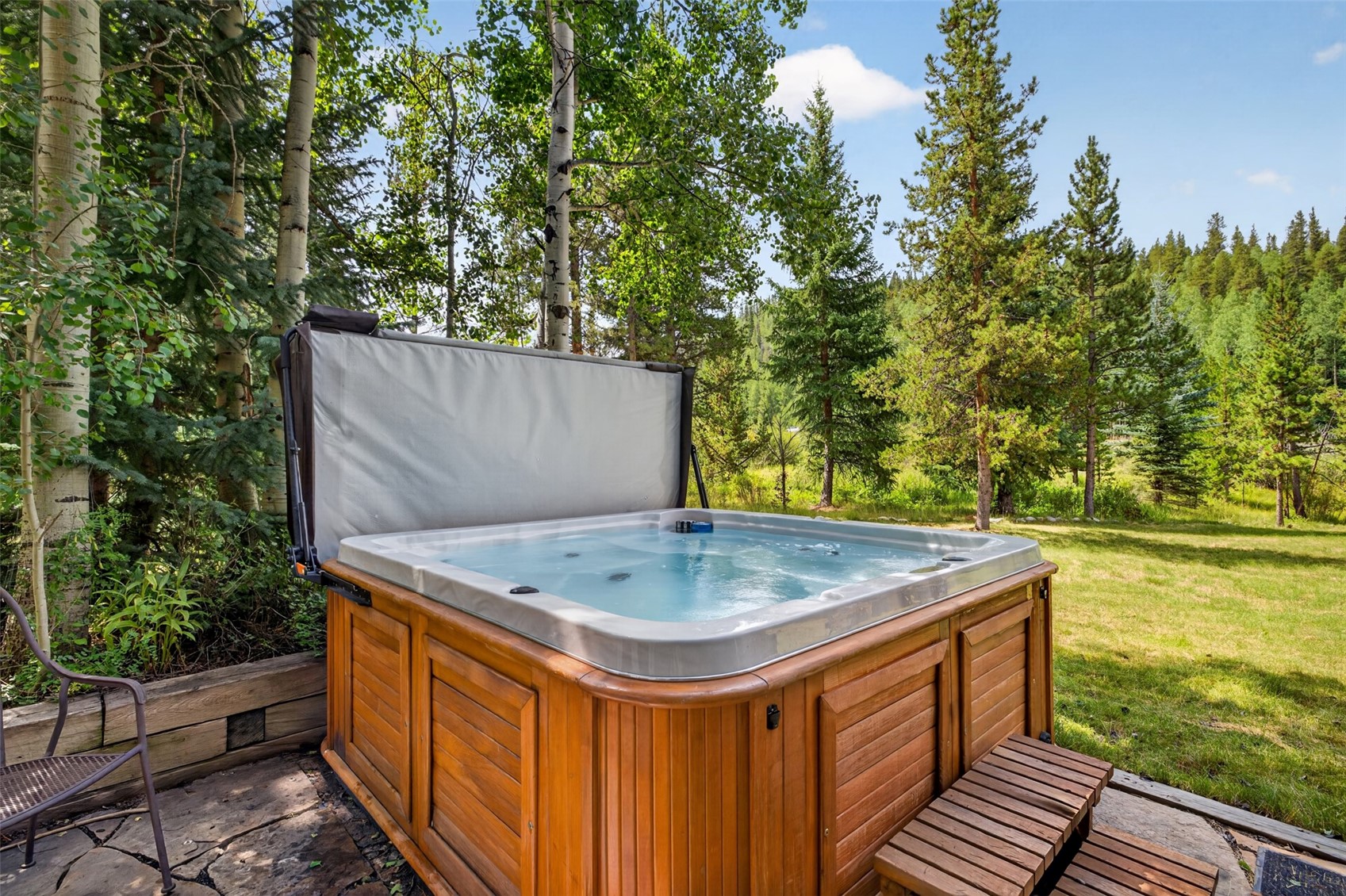 A wooden-paneled hot tub sits on a stone patio, featuring an open cover and clear, bubbling water. The spa is nestled in a lush, natural setting surrounded by tall aspen and pine trees, with a grassy lawn extending into the background. The scene captures a serene, private outdoor retreat perfect for relaxation.