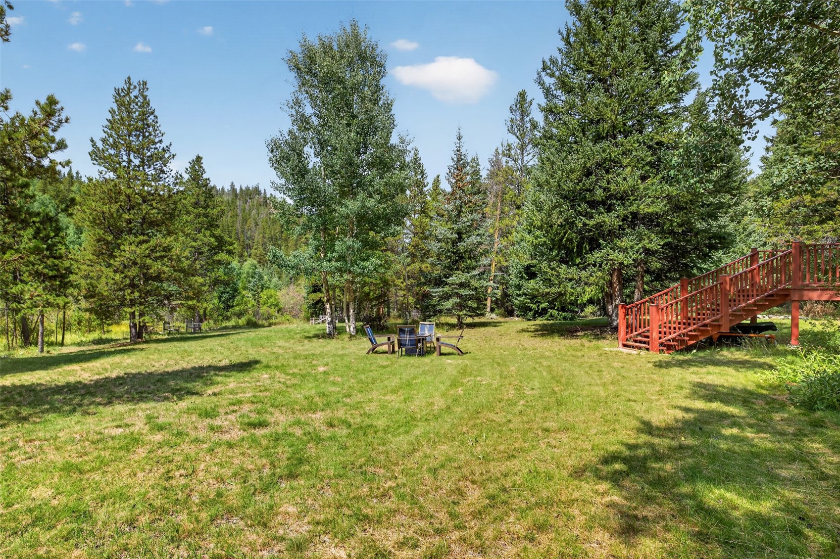 This expansive, sun-drenched backyard features a lush green lawn surrounded by a dense forest of tall pine and aspen trees. A set of wooden stairs leads up to a raised deck on the right, while a small seating area with outdoor chairs is positioned in the center of the yard. The scene captures a peaceful, private outdoor retreat with a natural, wooded landscape.