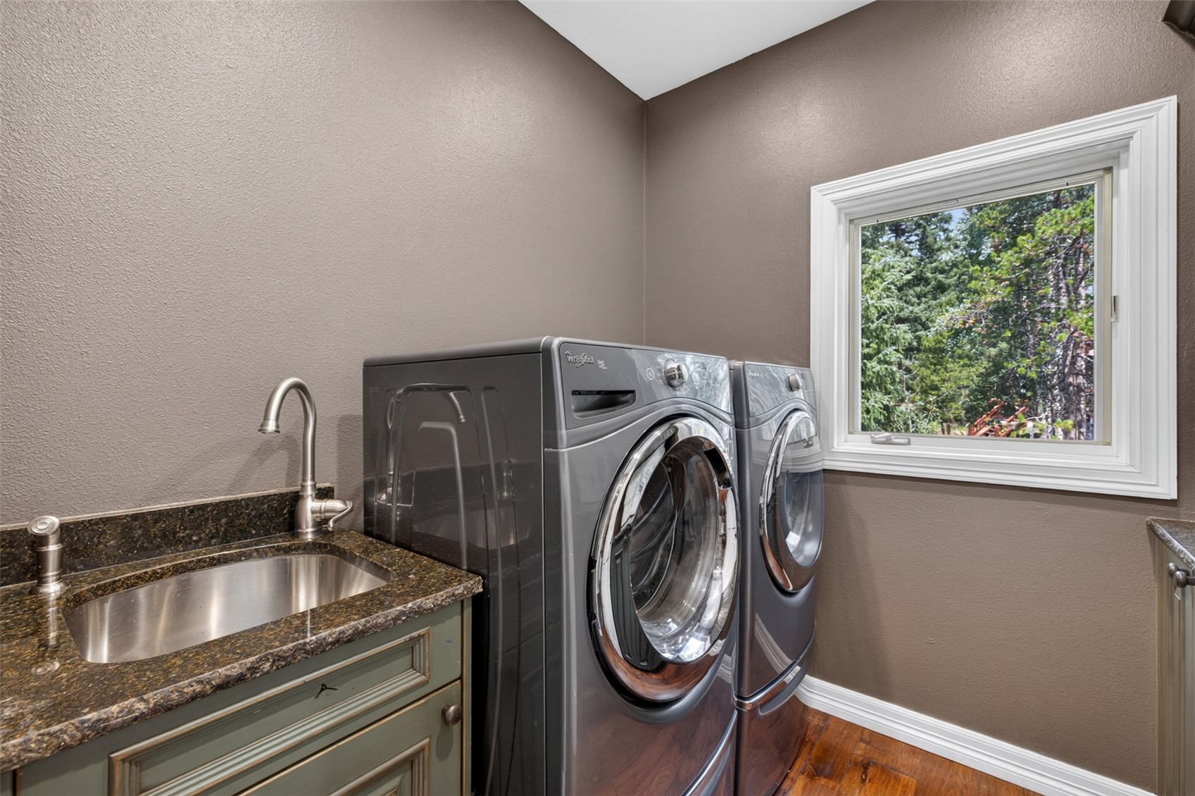 This functional laundry room features a pair of dark gray, front-loading washer and dryer units positioned next to a built-in sink cabinet. The space is accented by a dark stone countertop, a modern faucet, and a window that provides a view of the surrounding trees. The room is finished with dark-toned walls and warm wood flooring, creating a clean and practical utility area.