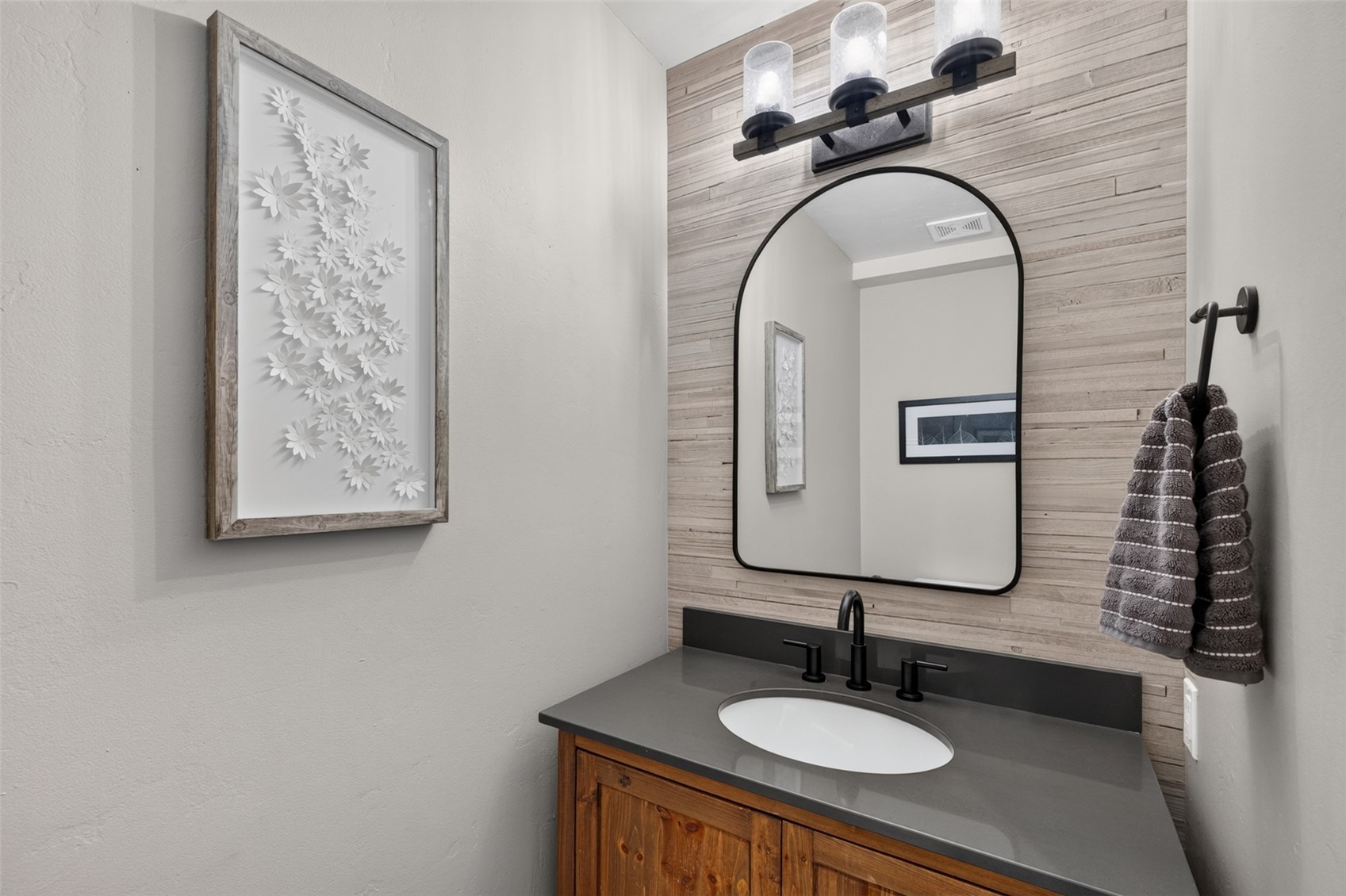 This modern powder room features a rustic wood vanity topped with a dark, solid-surface countertop and a matte black faucet. The wall behind the vanity is accented with horizontal wood-look paneling, complemented by an arched mirror and a three-light industrial-style fixture. A framed floral art piece hangs on the adjacent wall, and a grey towel hangs from a circular wall-mounted ring, creating a cohesive and stylish aesthetic.