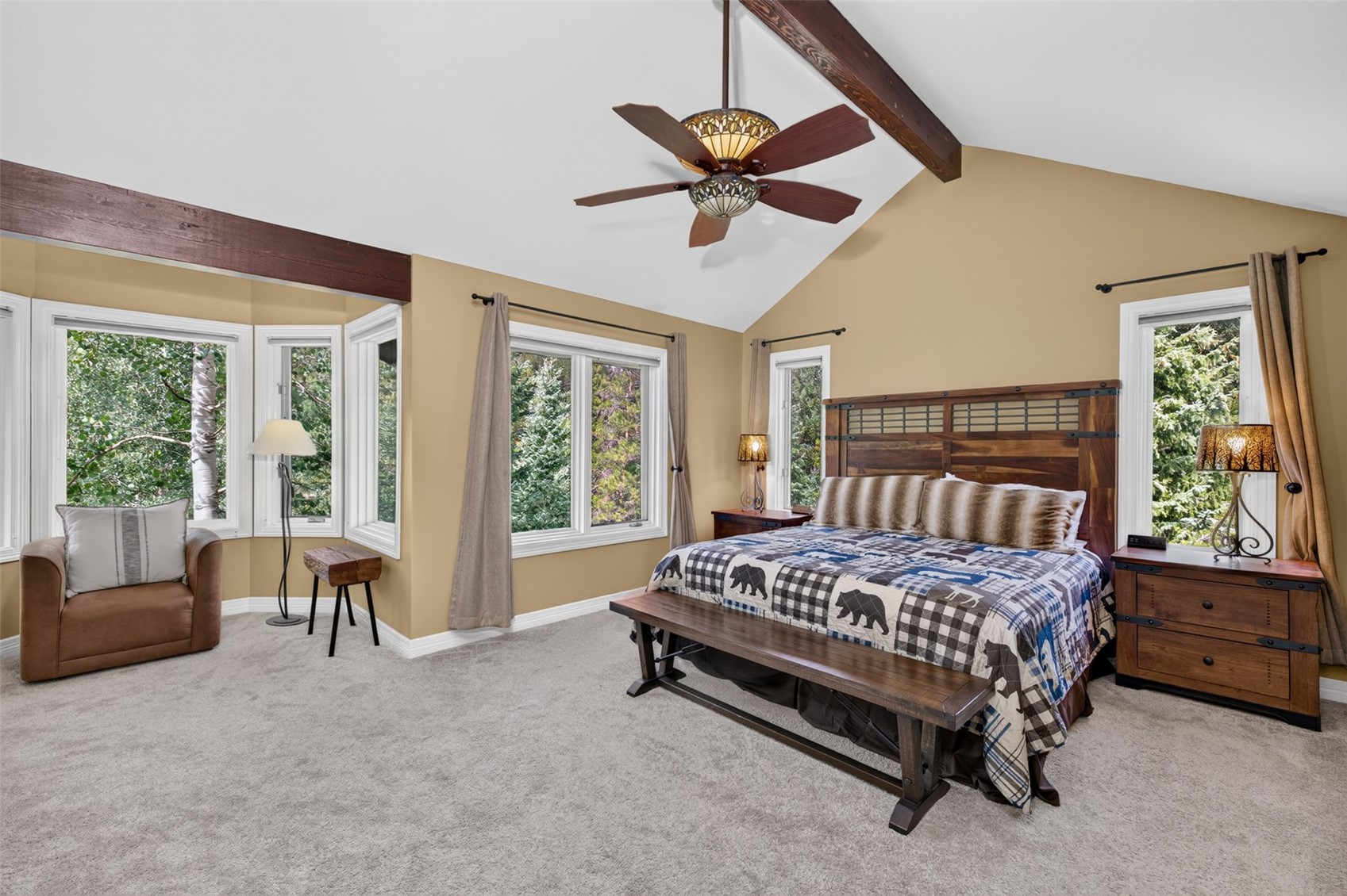 This spacious primary bedroom features a rustic, lodge-inspired aesthetic with a vaulted ceiling accented by dark wood beams. The room is furnished with a large wooden bed frame, a matching bench, and a cozy armchair tucked into a bay window nook. Large windows provide ample natural light and views of the surrounding trees, while a decorative ceiling fan with integrated lighting serves as a central focal point.