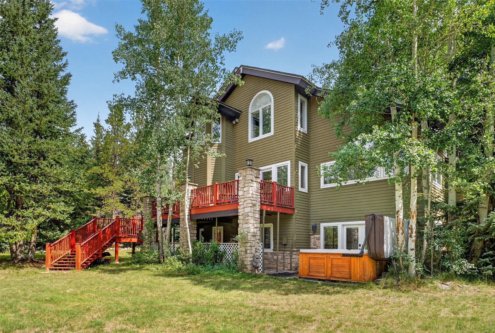 This rear view of a multi-story home showcases olive-green siding and a prominent elevated deck with red railings, supported by stone-clad pillars. A wooden hot tub sits on the ground level near the base of the house, surrounded by mature aspen and pine trees. The scene captures a peaceful, wooded backyard setting with a wooden staircase leading up to the main deck area.