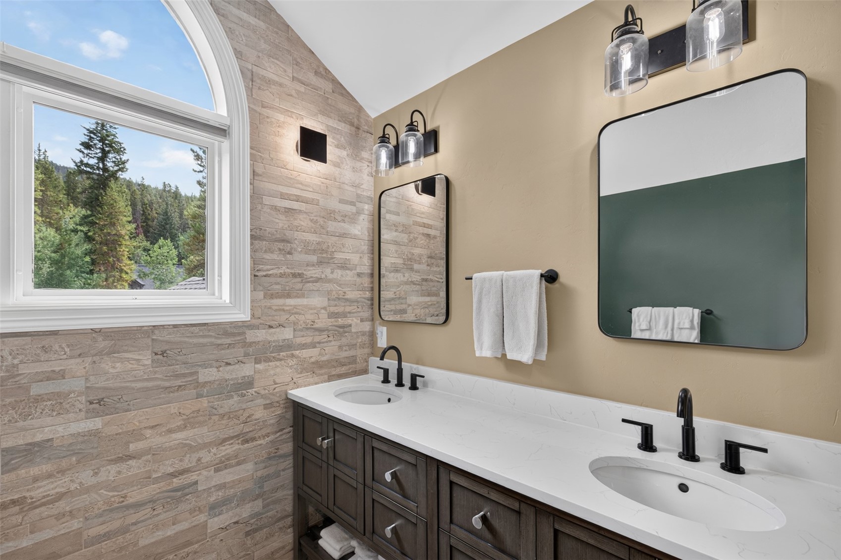 This primary bathroom features a double vanity with a white countertop and dark wood cabinetry, complemented by matte black fixtures. A striking stone-tiled accent wall adds texture, while a large arched window provides natural light and a scenic view of the surrounding forest. The space is finished with two rectangular mirrors and modern industrial-style vanity lighting, creating a sophisticated and serene atmosphere.