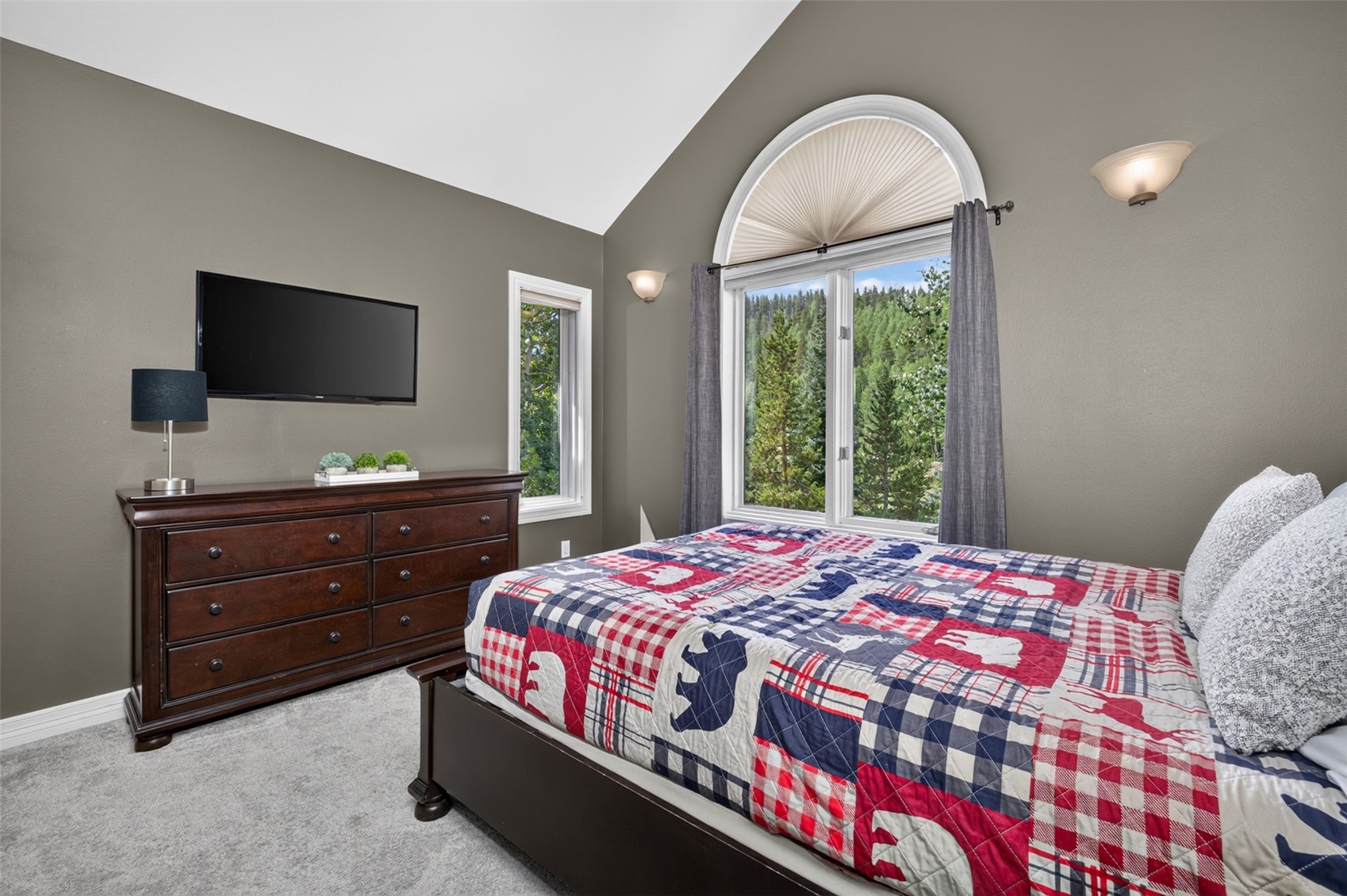 This cozy bedroom features a vaulted ceiling and a large arched window that frames a scenic view of the surrounding forest. The room is furnished with a dark wood dresser topped with a television and a bed dressed in a rustic, plaid-patterned quilt featuring bear motifs. The neutral wall color and soft carpeting create a comfortable, mountain-inspired retreat.