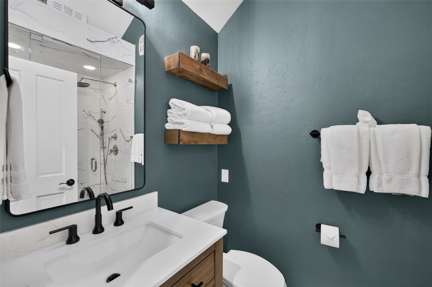 This modern bathroom features a vanity with a white countertop and dark hardware, complemented by a large, black-framed mirror. The walls are painted a deep, moody teal, providing a striking contrast to the white towels and the light wood floating shelves. A glass-enclosed shower with patterned stone walls is visible in the reflection, creating a cohesive and stylish aesthetic.