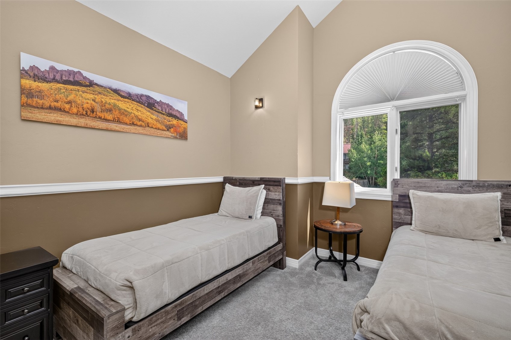 This guest bedroom features two twin beds with rustic, weathered-wood headboards and neutral bedding. The room is accented by a two-tone wall treatment with a chair rail, a large arched window that brings in natural light, and a landscape art piece above one of the beds. A small round side table with a lamp sits between the beds, creating a cozy and inviting atmosphere.