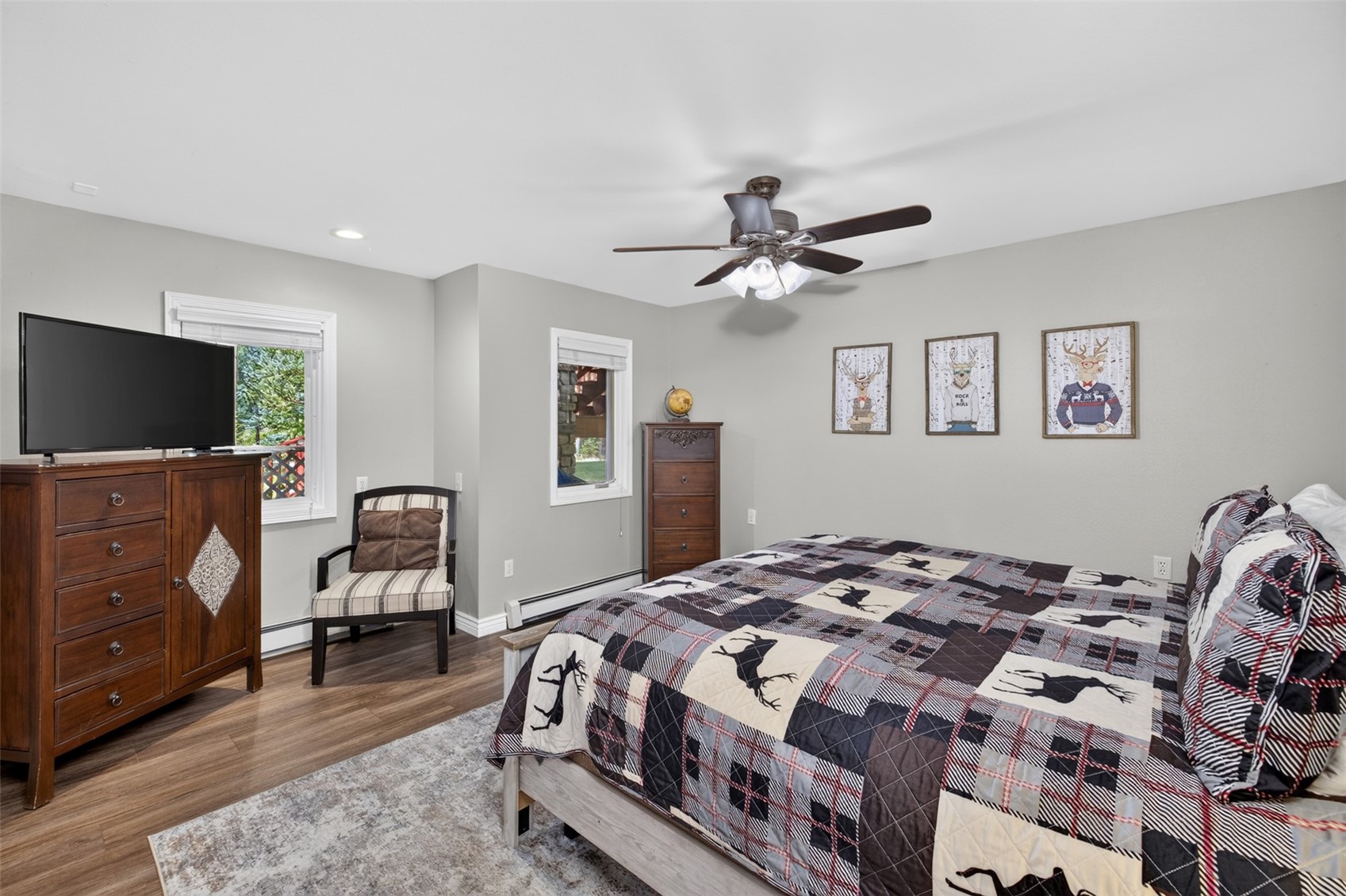 This cozy bedroom features a rustic-themed aesthetic with a plaid quilt adorned with deer silhouettes and matching pillows. The room is furnished with a dark wood dresser topped with a television, a tall matching chest of drawers, and a striped accent chair positioned near a window. Three framed prints of stylized deer hang on the light gray wall above the bed, creating a cohesive and inviting mountain-lodge atmosphere.