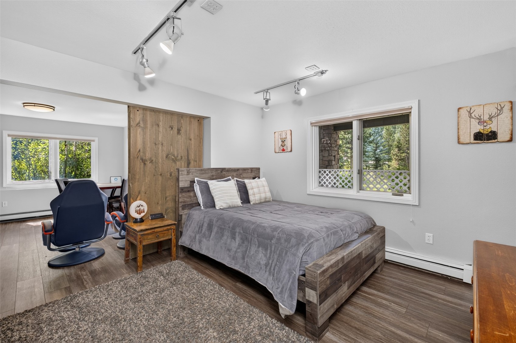 This bedroom features a rustic aesthetic with a wood-paneled headboard and matching bed frame, complemented by a gray comforter and patterned throw pillows. A sliding barn door separates the sleeping area from an adjacent office space, which contains a modern blue chair and a desk. The room is illuminated by track lighting and natural light from a window, creating a cozy and functional living environment.