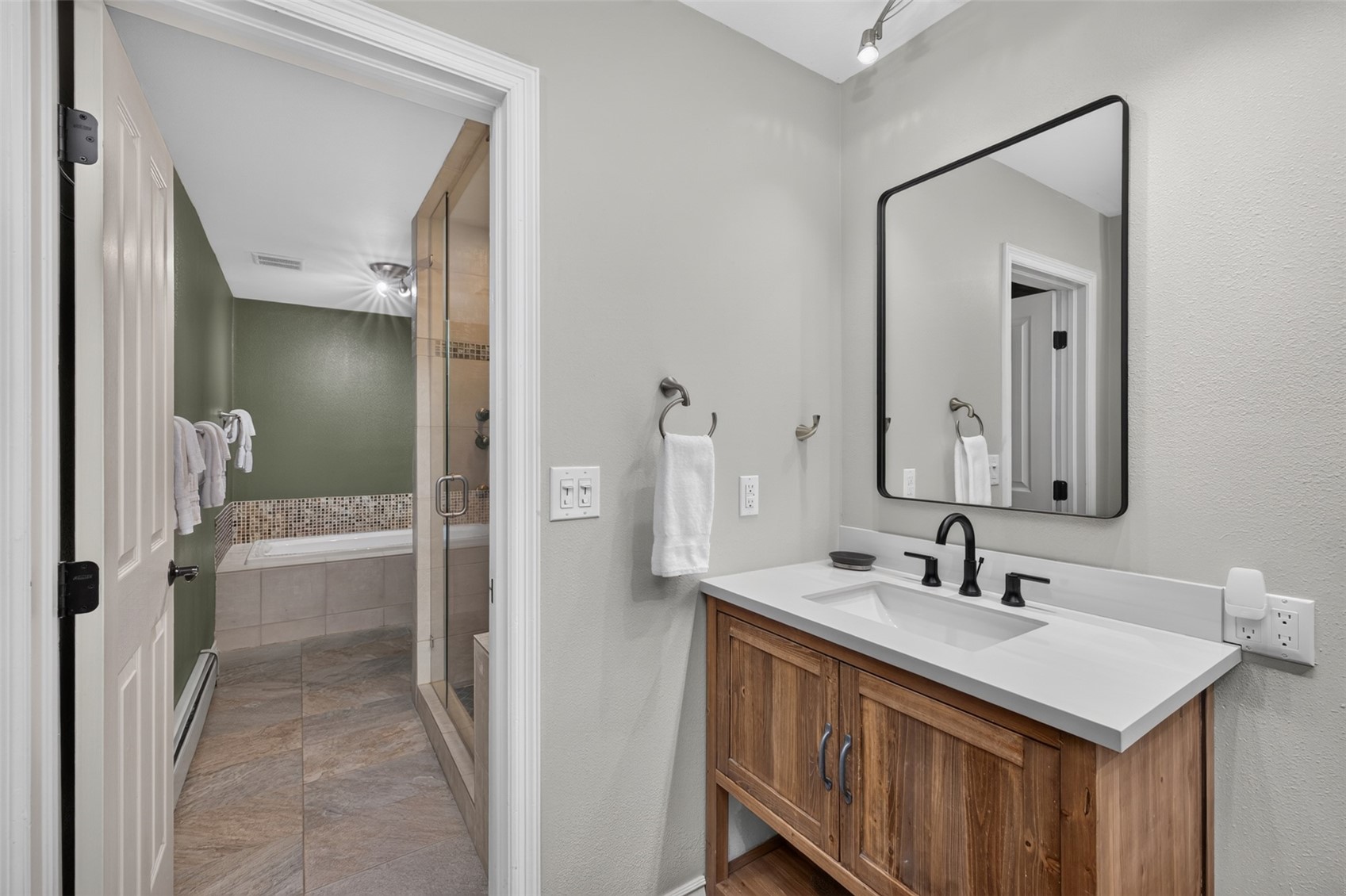 This primary bathroom features a rustic-style wooden vanity with a light-colored countertop and a modern black-framed mirror. The space opens into a secondary area containing a glass-enclosed shower and a soaking tub set against a green wall with decorative tile accents. The neutral color palette and clean lines create a functional and inviting atmosphere.