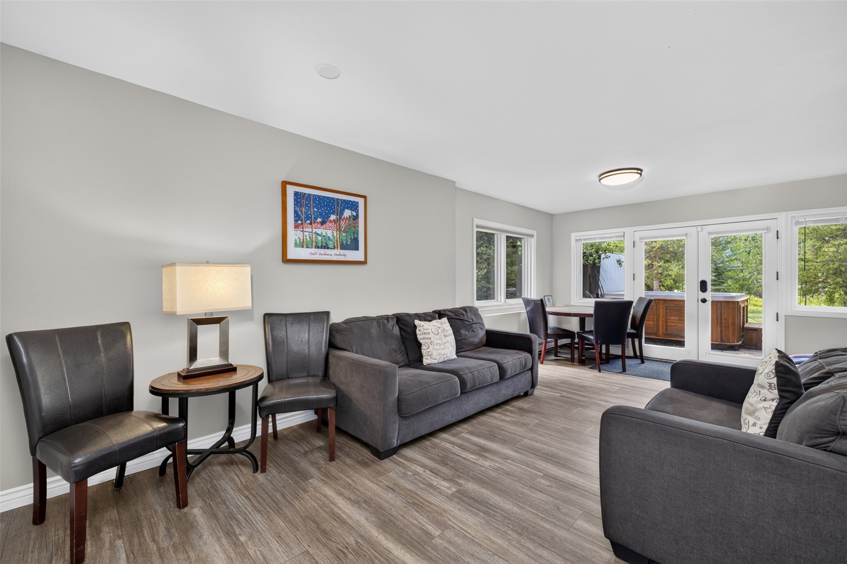 This open-concept living area features light wood-look flooring and neutral gray walls, creating a bright and inviting atmosphere. The space is furnished with two dark gray sofas, a small round side table with a lamp, and a dining set positioned near a set of glass doors that lead to an outdoor deck with a hot tub. A framed piece of art hangs on the wall, adding a touch of color to the otherwise minimalist and functional design.