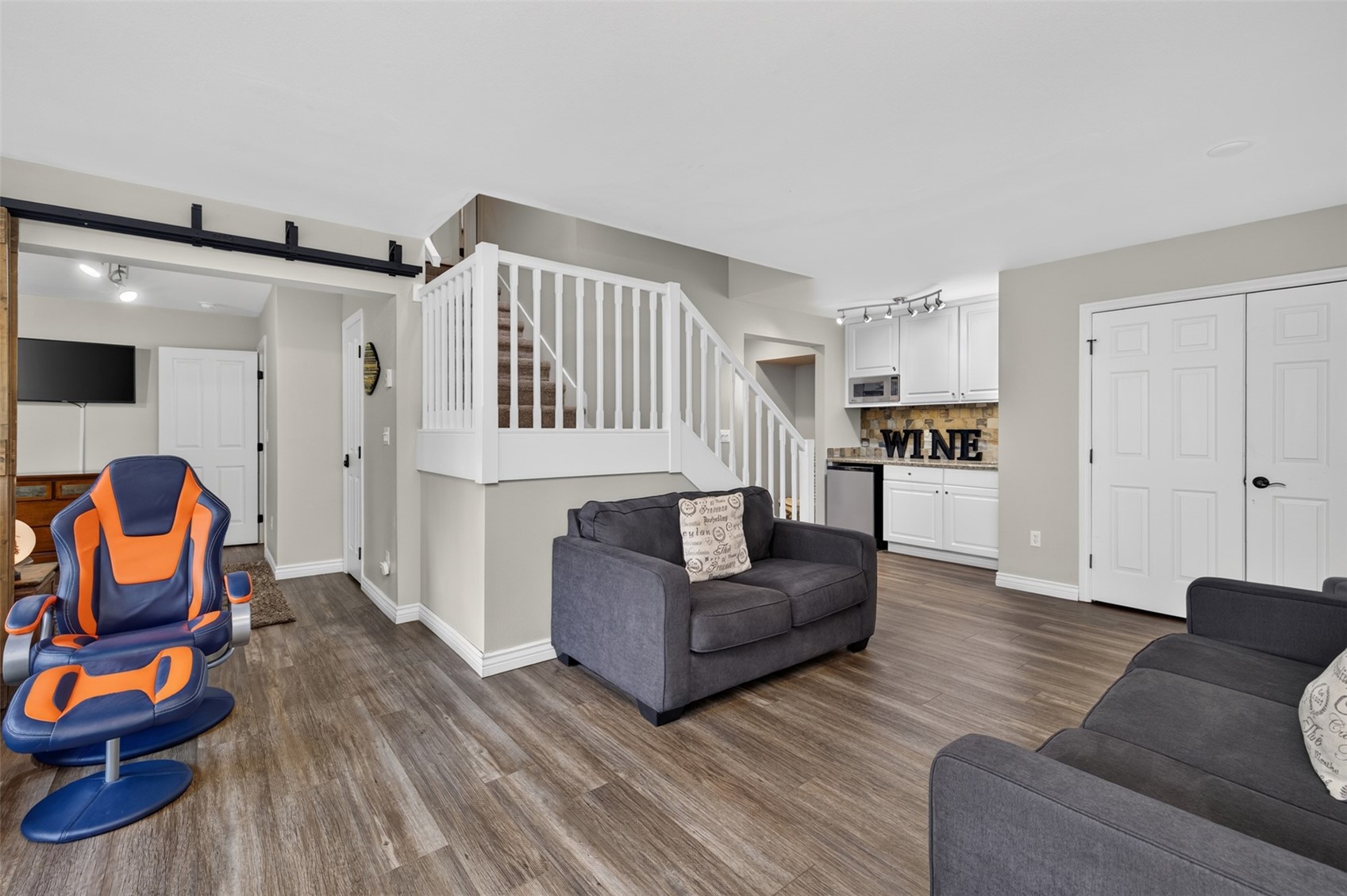 This interior view captures a versatile living area featuring a dark gray loveseat and a matching sofa, complemented by a modern blue and orange gaming chair. The space includes a small wet bar area with white cabinetry and a stone-tiled backsplash, situated near a staircase with white railings. The room is finished with light-toned wood-look flooring and neutral wall colors, creating a functional and inviting multi-purpose living space.