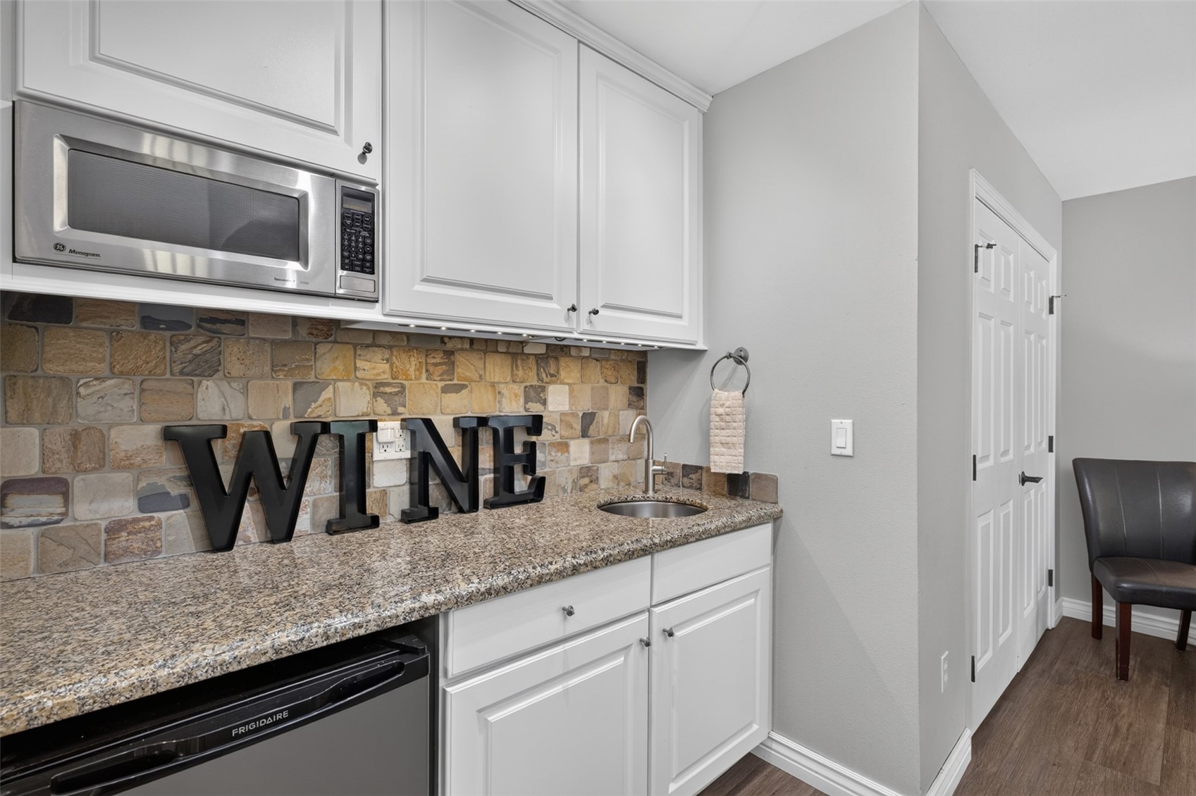 This compact wet bar area features white cabinetry, a granite countertop, and a stone-tiled backsplash. A stainless steel microwave is mounted above the counter, which also includes a small sink and a Frigidaire under-counter appliance. The space is accented by large decorative letters spelling 'WINE' and is situated next to a doorway leading to a room with a single dark chair.