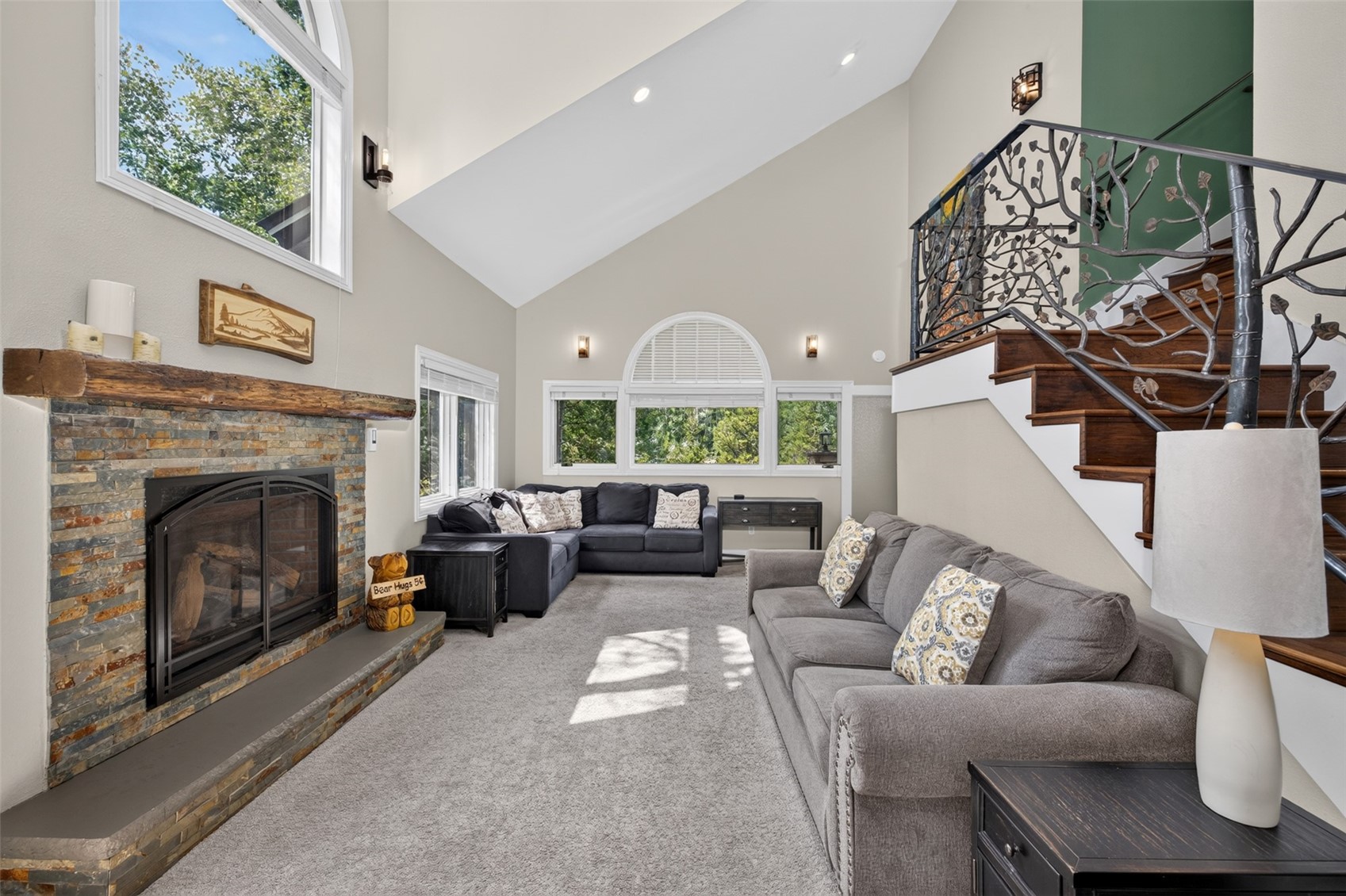 This spacious living room features a vaulted ceiling and a prominent stone-clad chimney breast with a rustic wood mantel. The room is furnished with two large grey sofas and includes a staircase with decorative metal railings leading to an upper level. Large windows, including an arched window, allow natural light to fill the space, creating an open and inviting atmosphere.